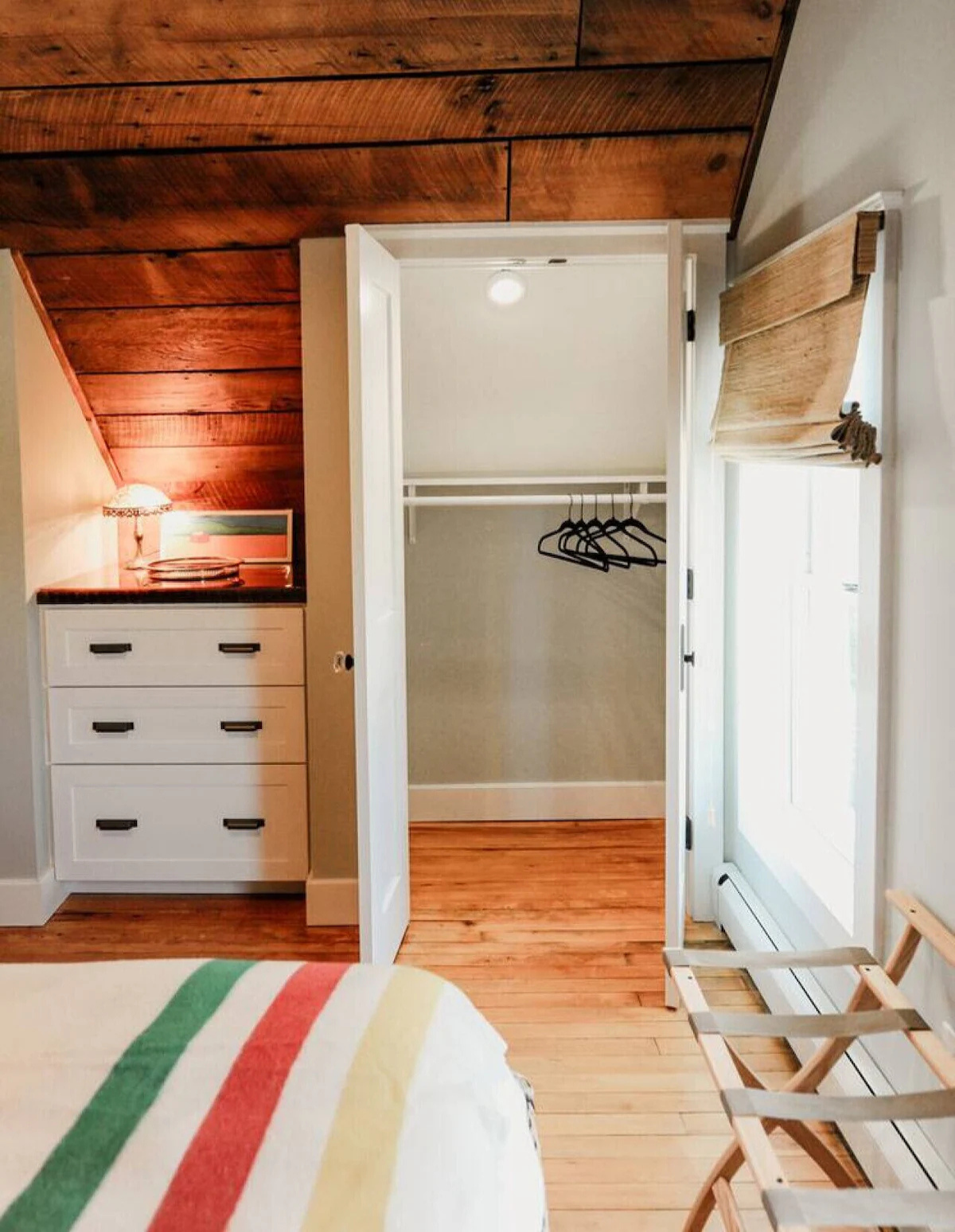 Open closet with hanging rods and four black hangers, room with wood floor, wall-mounted heater, and window with beige Roman shade, adjacent to ceiling with wooden panels and a dresser with a lamp and artwork.