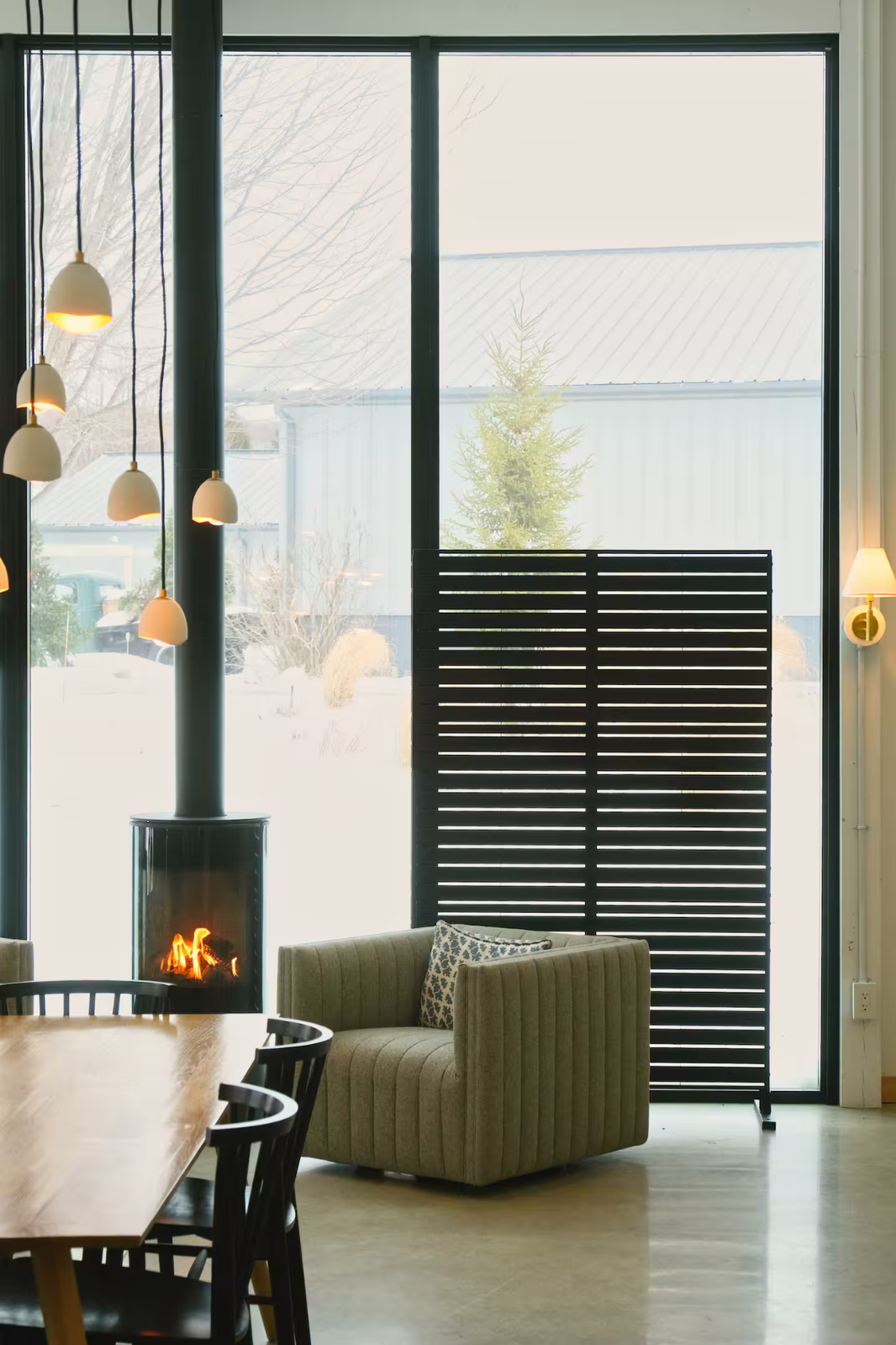 Interior of a cozy living space with a wood-burning stove, beige armchair with a patterned pillow, a wooden dining table with black chairs, large floor-to-ceiling window, and modern lighting fixtures.