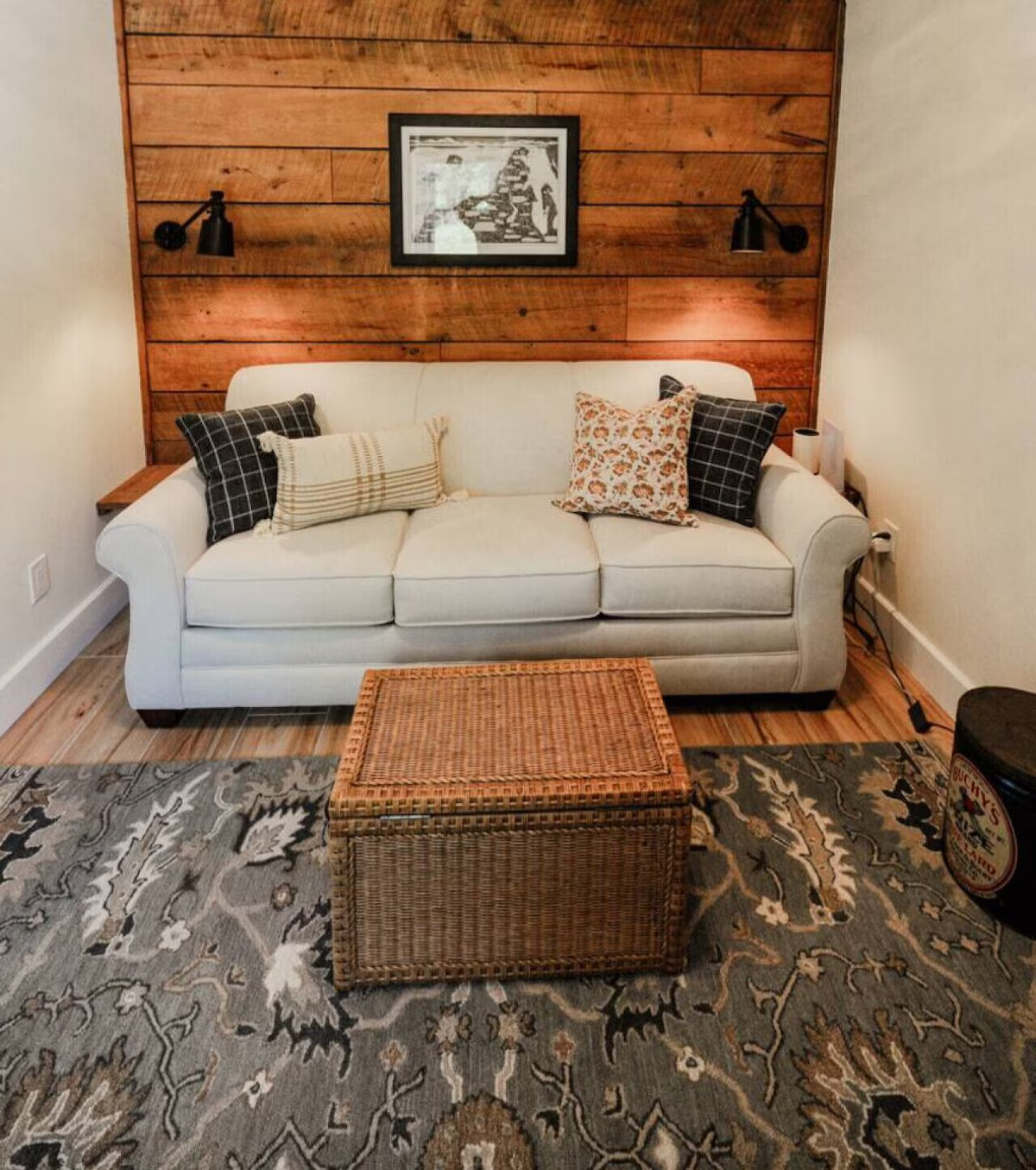 A cozy living room with a white sofa, decorative pillows, a wooden wall, framed artwork, two black wall-mounted reading lights, a wicker coffee table, a patterned area rug, and a drum-shaped side table.