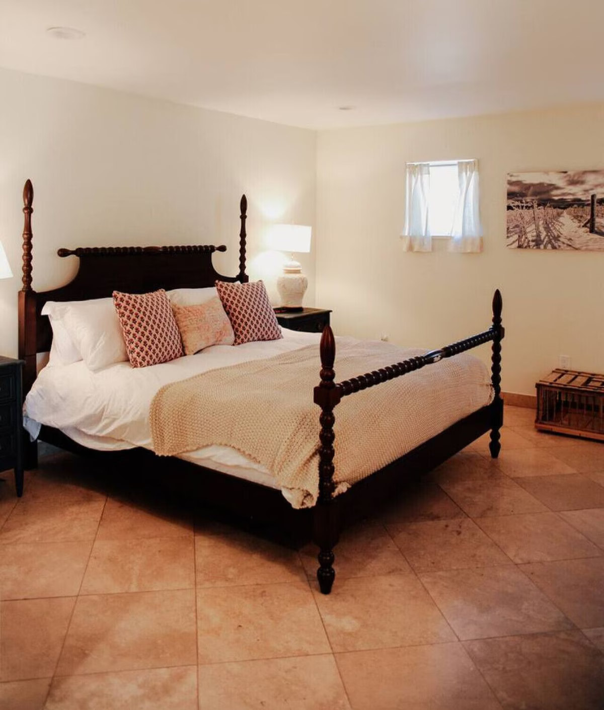 A bedroom with a dark wooden four-poster bed, white bedding, and decorative pillows. There are two nightstands with lamps, a window with white curtains, and a wall art piece of a snow-covered landscape.