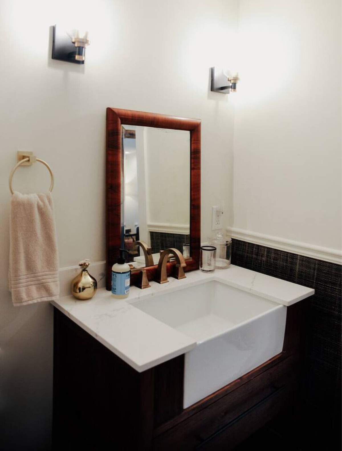 Bathroom vanity with a white rectangular sink, a wooden framed mirror, a chrome faucet, a gold vase, hand soap, a towel holder with a beige towel, and a few glass containers. Two wall-mounted lights illuminate the area.