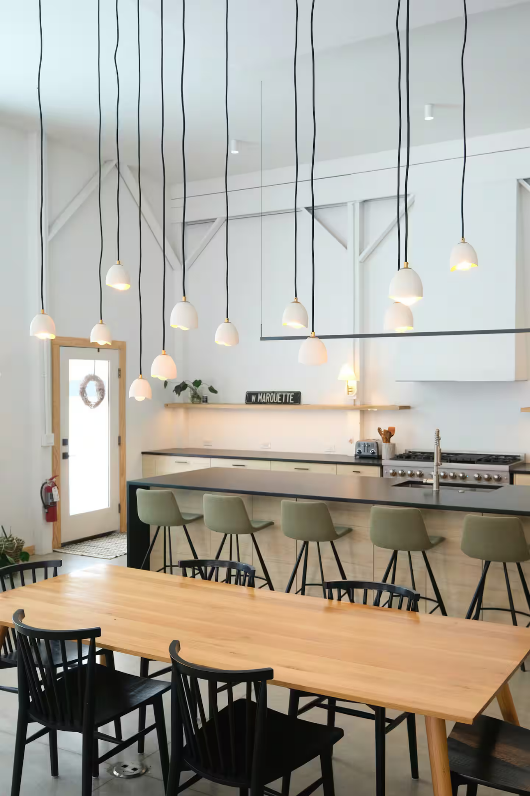 Modern kitchen with a large wooden dining table, black chairs, green bar stools, and unique pendant lighting fixtures hanging from the ceiling.