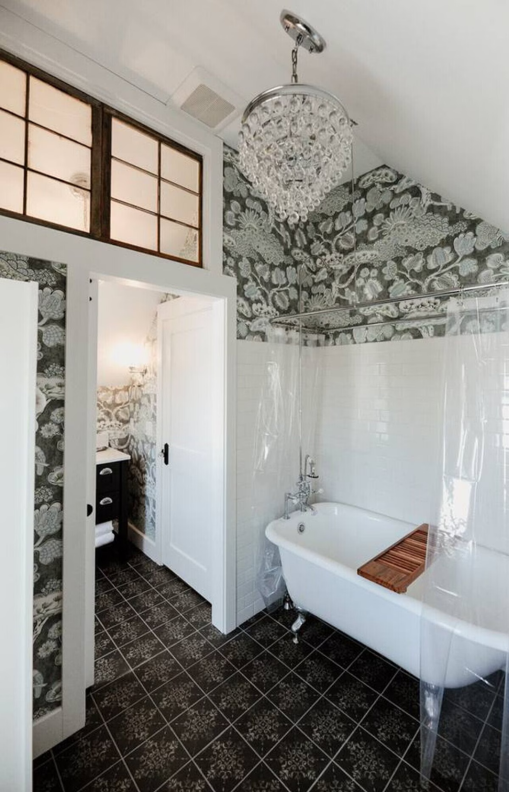 Bathroom with black and white floral wallpaper, a vintage clawfoot bathtub with a wooden bath tray, a black and white tiled floor, a chandelier hanging from the ceiling, and a small black vanity with white towels in a separate room visible through a 