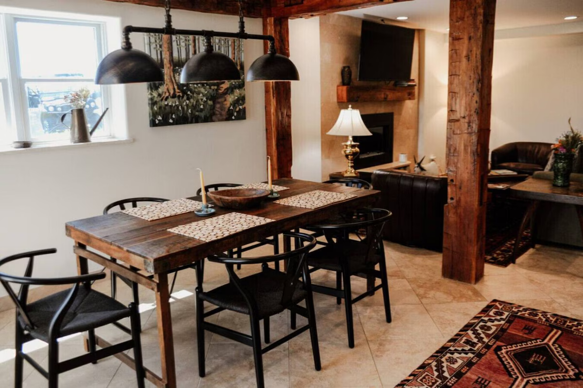 Dining room with a rustic wooden table, six black chairs, black pendant lights, a window with a watering can and vase on the sill, wall art, a fireplace with a television above it, and a cozy seating area with a lamp and a coffee table.