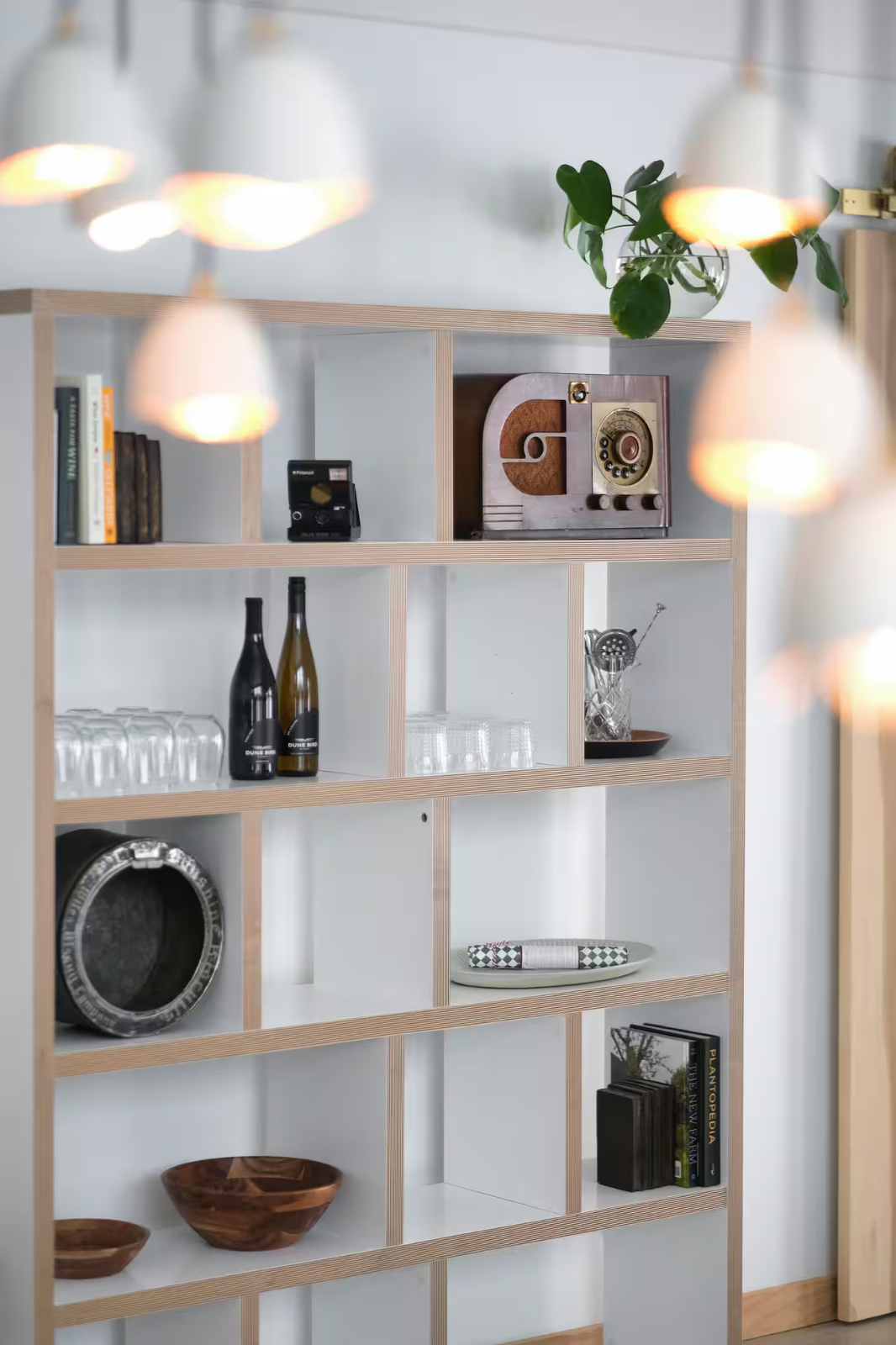 Open wooden shelving unit with decorative items, glassware, books, and a vintage radio, with a potted plant on top, in a modern interior.