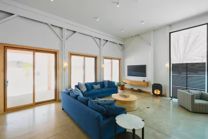 Modern living room with large blue sectional sofa, a round coffee table, and a smaller side table. Features large sliding glass doors, a wall-mounted TV, and a fireplace. Bright, spacious, and minimalistic decor.