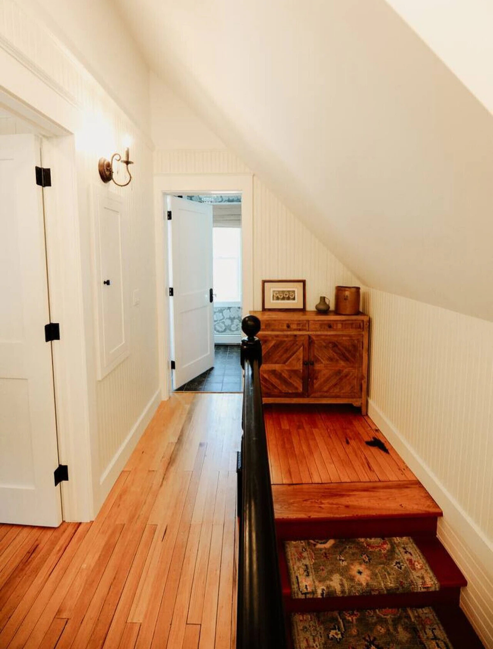 A cozy hallway with wooden floors, a staircase leading down with a decorative carpet, a wooden sideboard with framed pictures and vases, and a door that opens to a bathroom with a white tub and patterned tiles.
