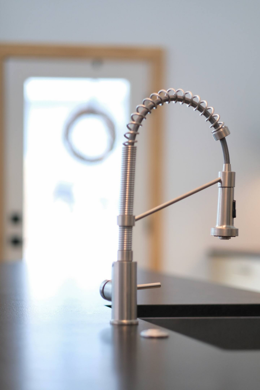 Modern stainless steel kitchen sink faucet with a flexible spring spout, installed on black countertop in a kitchen with natural light coming in through a door with a wreath hanging outside.