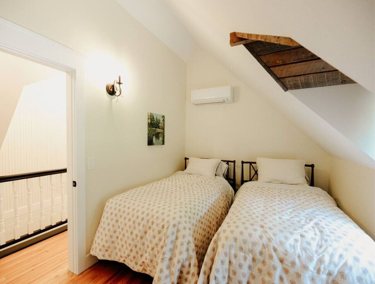 Two twin beds with beige patterned bedspreads in an attic bedroom with sloped ceiling, a small landscape painting on the wall, a wall-mounted air conditioner, a rustic wooden panel on the ceiling, and a wall sconce light.
