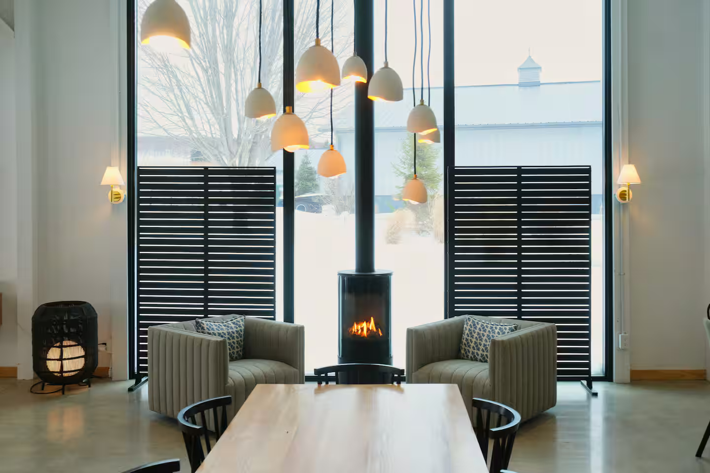 Modern living room with large window, fireplace, beige armchairs, a wooden table, and hanging pendant lights.