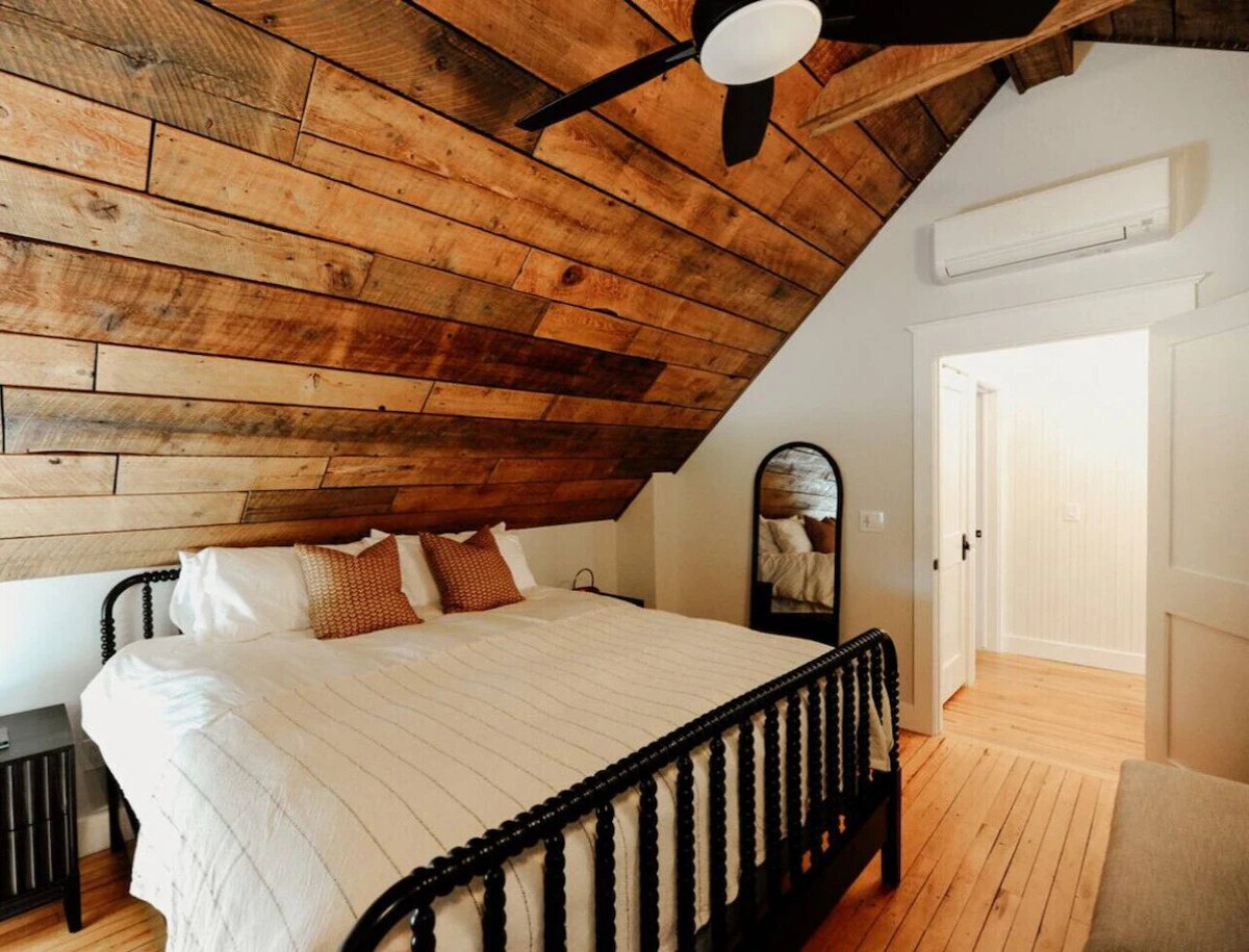 A bedroom with a slanted wooden accent wall, a black metal bed frame with white bedding, two brown pillows, a black bedside table, a full-length mirror, an air conditioning unit, and a ceiling fan with black blades.