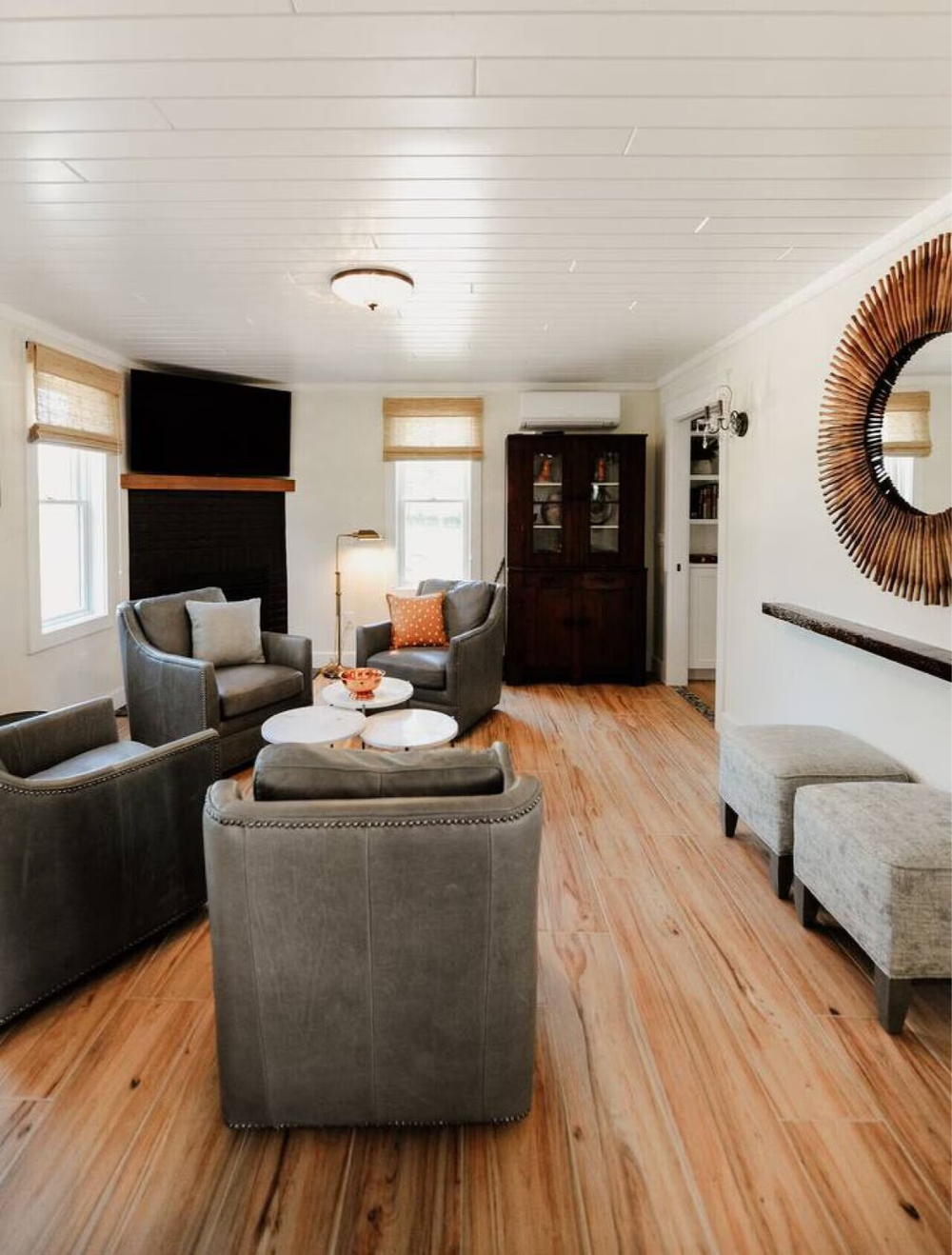 Living room with wooden flooring, gray armchairs, round white coffee tables, a dark wooden cabinet, large windows with blinds, a wall mirror, and a television mounted above a fireplace