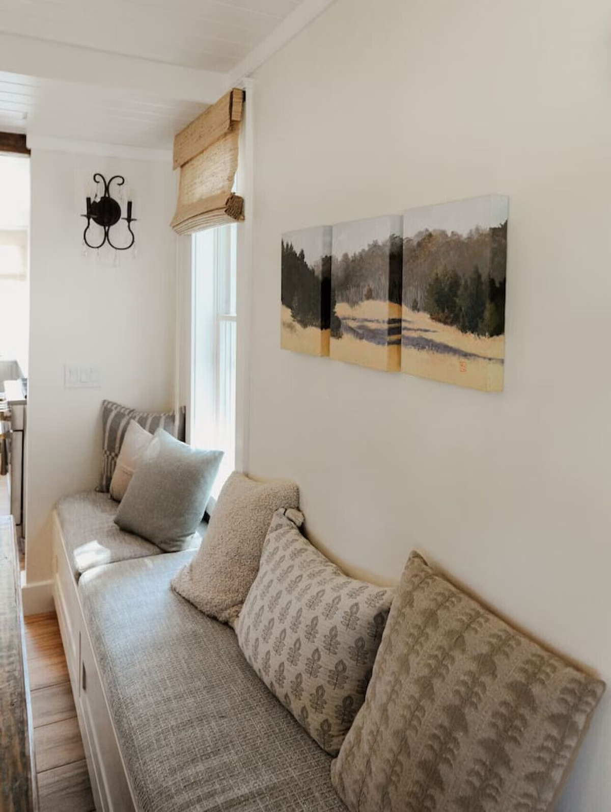 A cozy living room corner with a built-in bench seat decorated with various patterned and solid throw pillows. A wall-mounted triptych landscape painting depicts a wooded scene. There is a small window with a woven shade, and a black wall-mounted lig