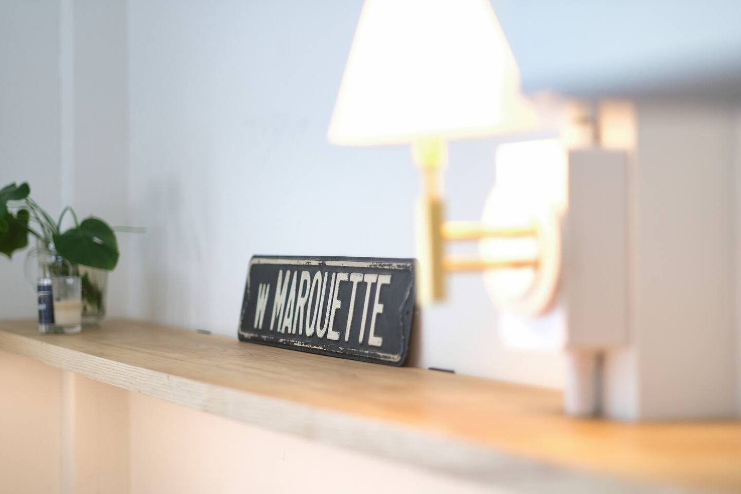 A wall-mounted lamp illuminating a wooden shelf with a black and white vintage street sign that reads 'W MARQUETTE', a small potted plant, and a candle.