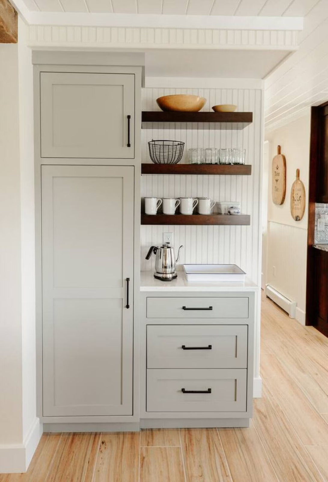 Kitchen cabinet with open shelves displaying bowls, a wire basket, glasses, mugs, and a teapot, in a home kitchen with light wood flooring.