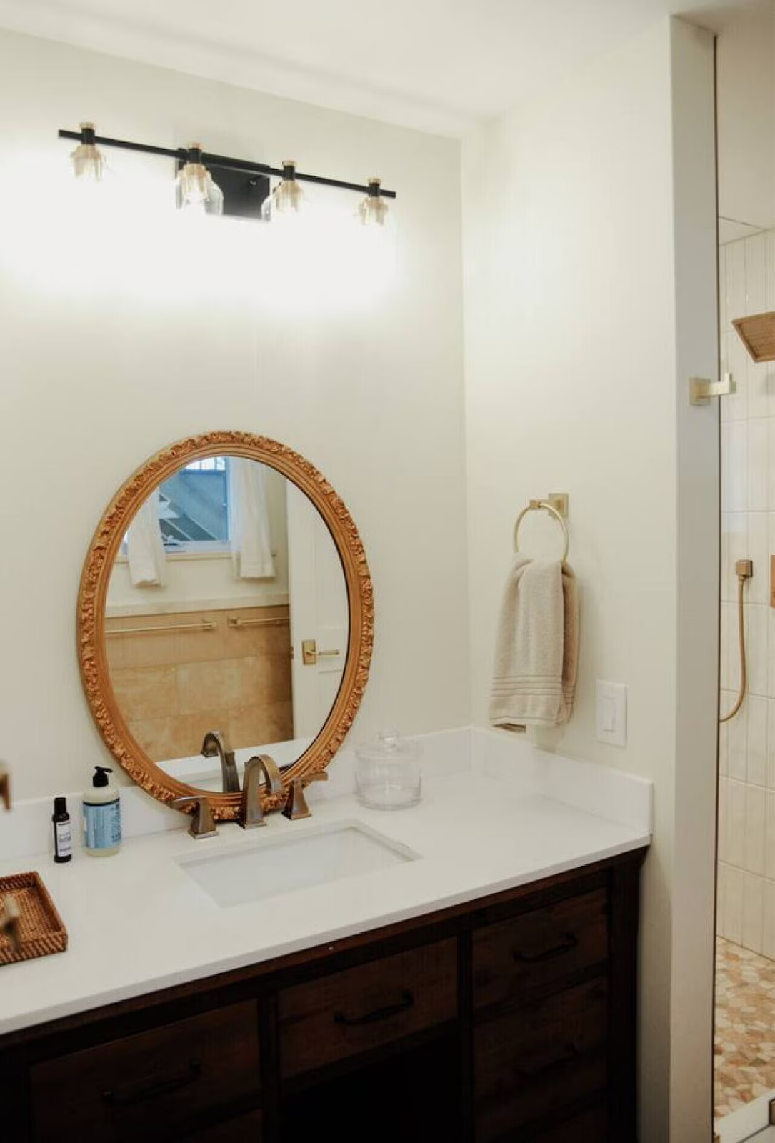 A bathroom vanity with a white countertop, oval ornate gold-framed mirror, a towel ring with a white towel, and a shower area with beige tiles visible through the doorway.