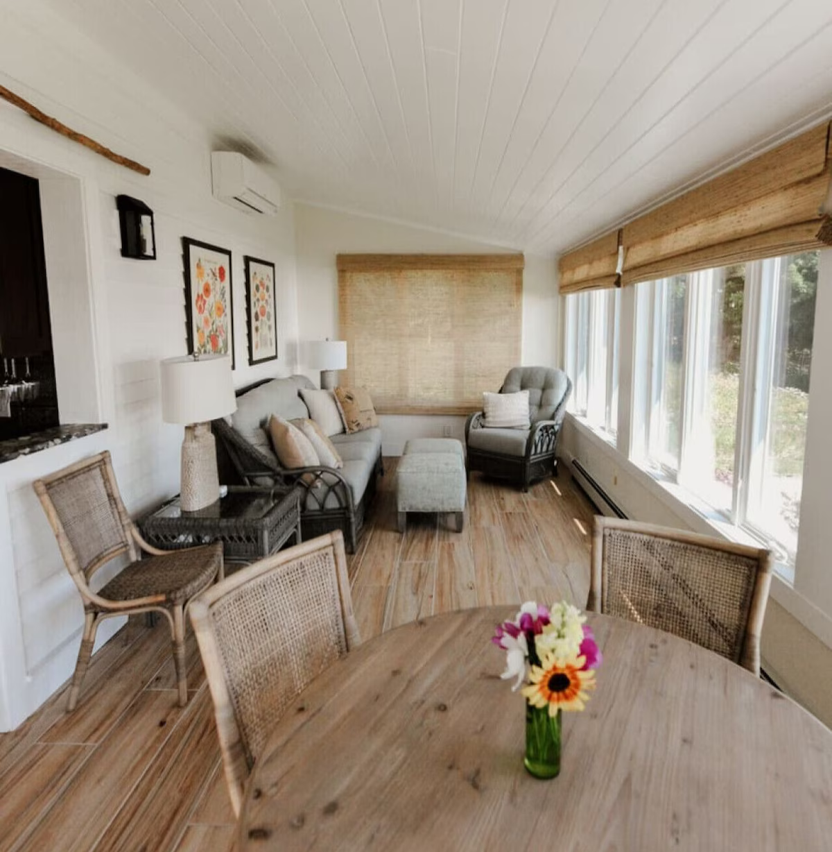 Cozy living room with wooden floors, large windows with bamboo shades, a gray sofa with beige pillows, a round table with a vase of colorful flowers, two wicker chairs, a gray armchair, and wall decorations.