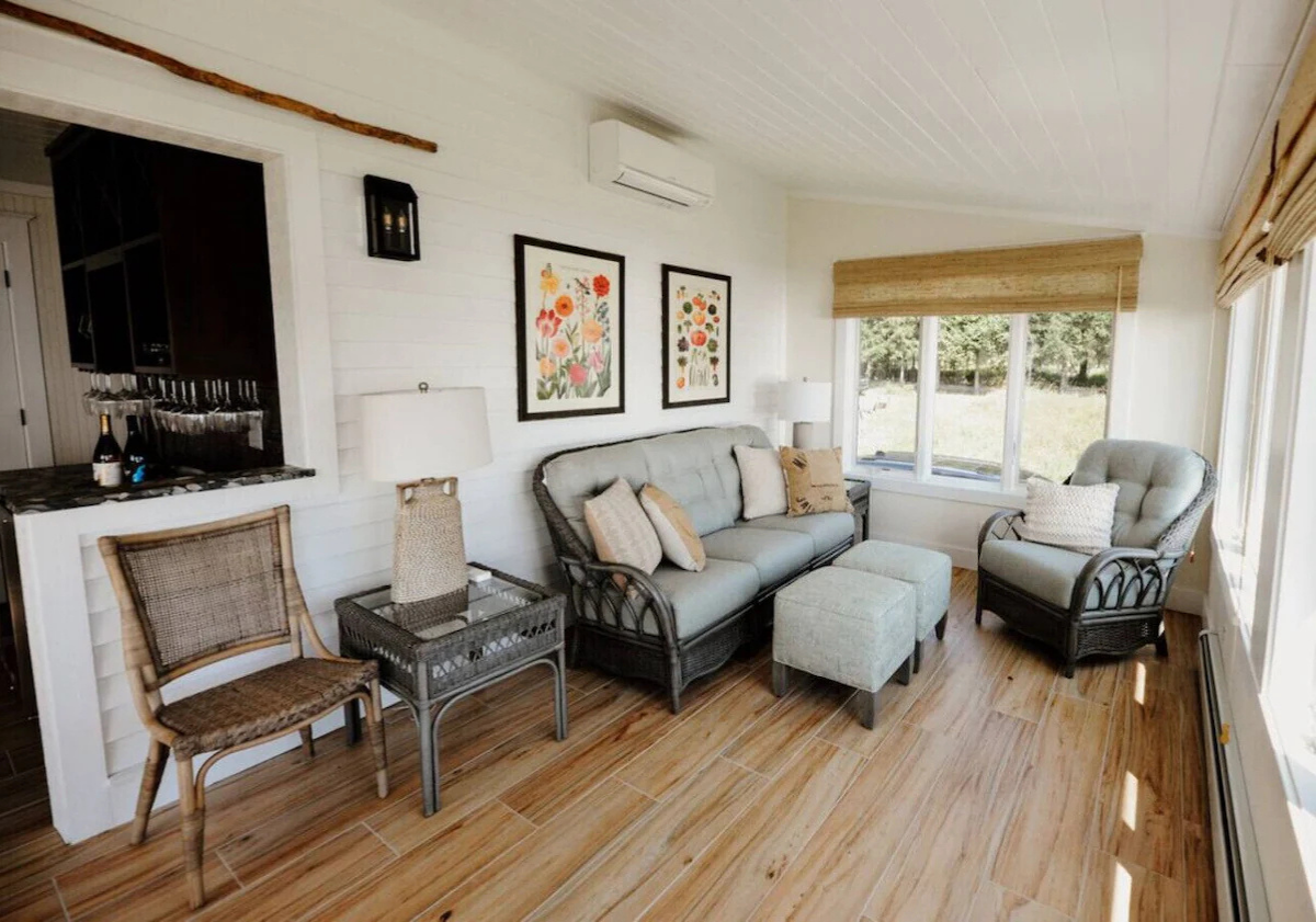 Coastal living room with wicker furniture, light-colored sofa and armchair, wooden flooring, large windows with shades, and framed floral artwork on white wall