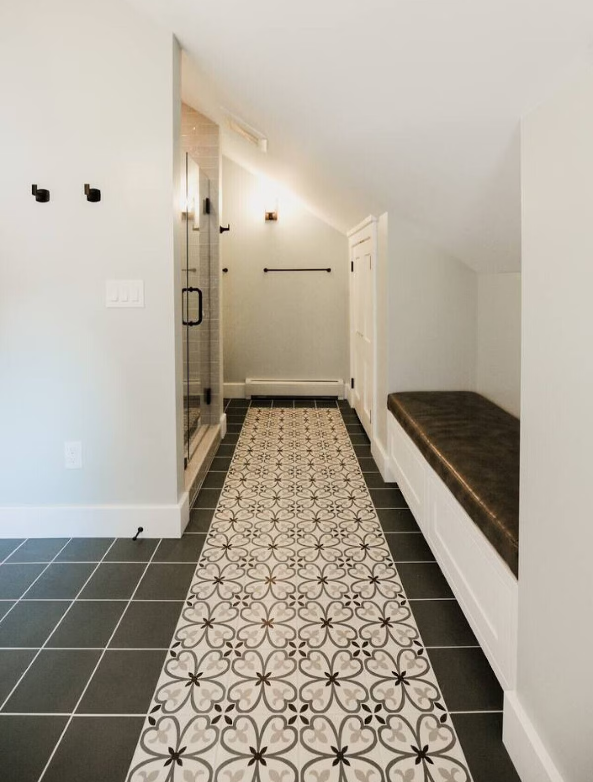 Bright entryway with patterned tile runner, black tiled floor, white walls, a built-in bench with dark cushion, and a glass door to a shower.