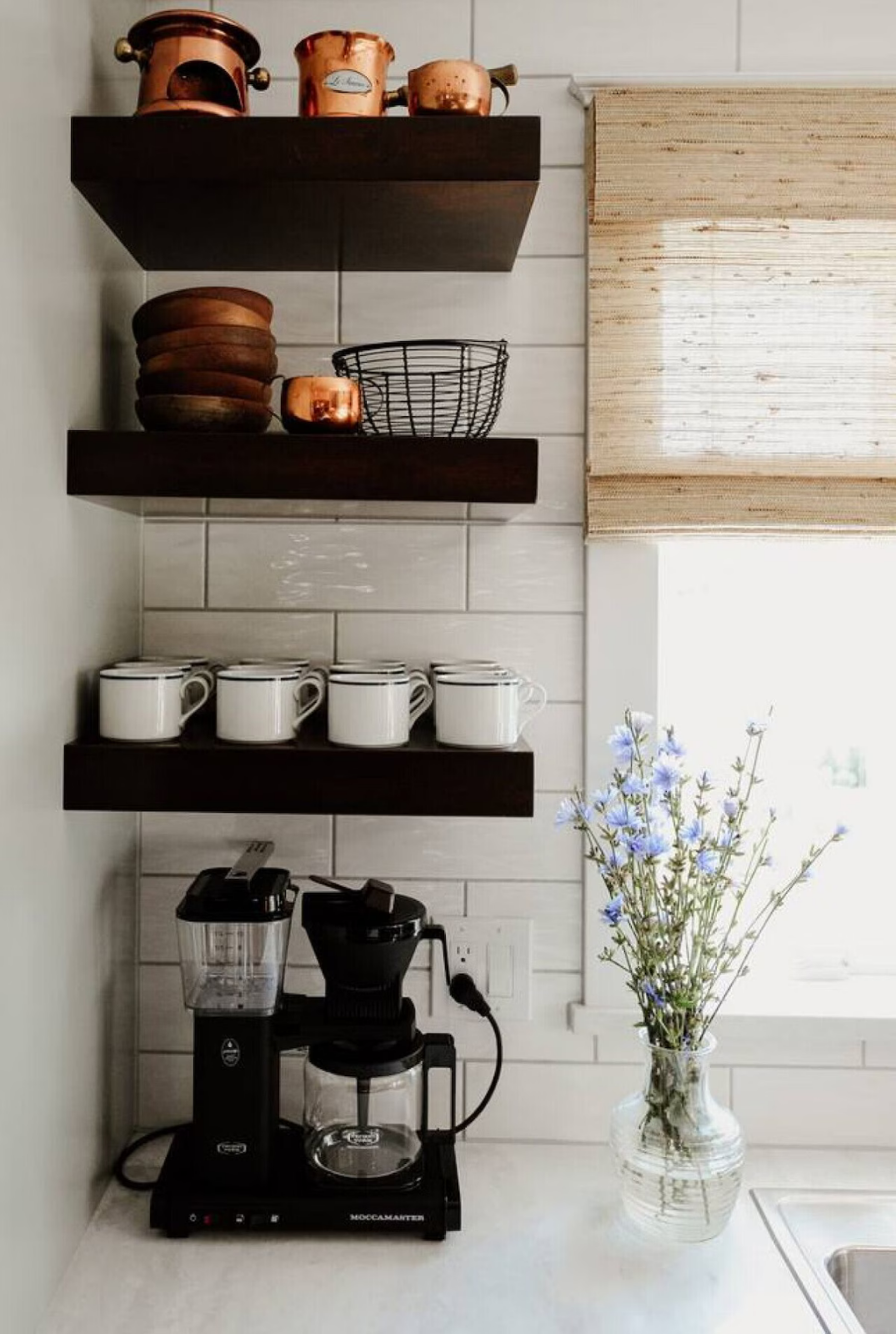 Kitchen countertop with black coffee maker, glass carafe, and coffee grinder next to a vase with blue flowers, with shelves holding copper pots, bowls, a wire basket, and white mugs.