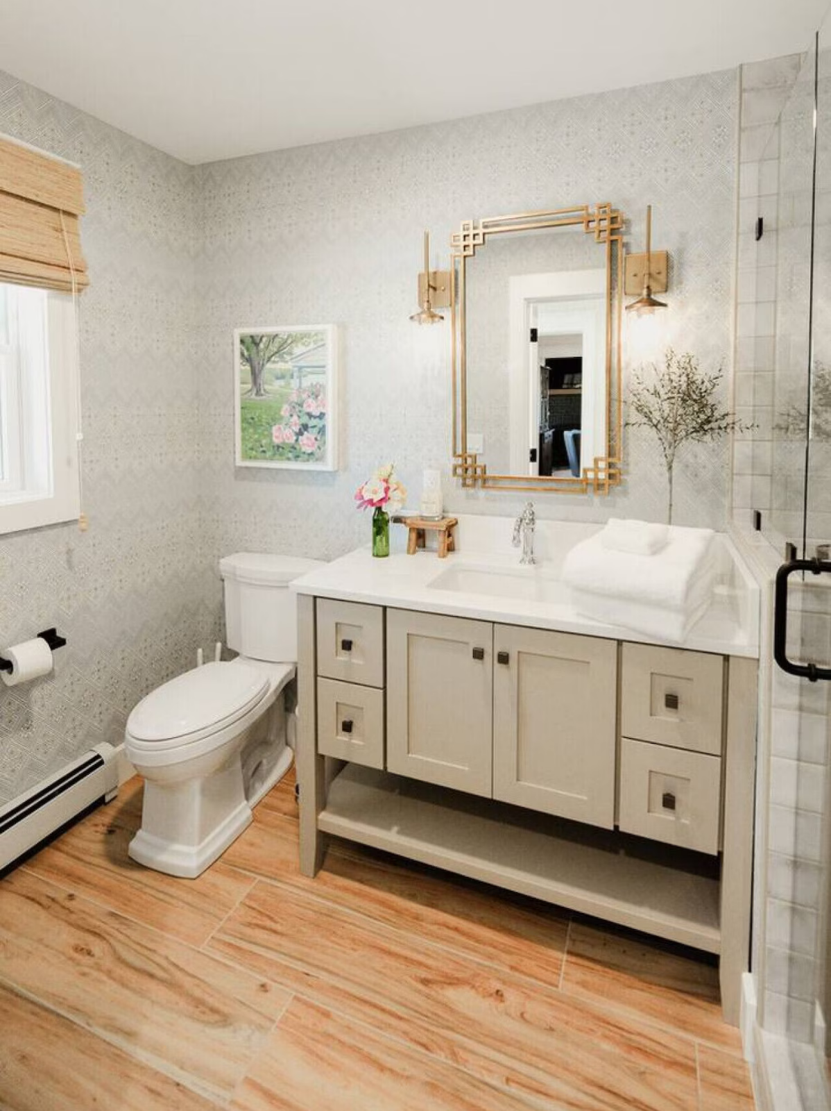 A bathroom with a wooden floor, a white toilet, a vanity with a sink, a gold-framed mirror, potted plants, a framed picture, and a window with bamboo blinds.