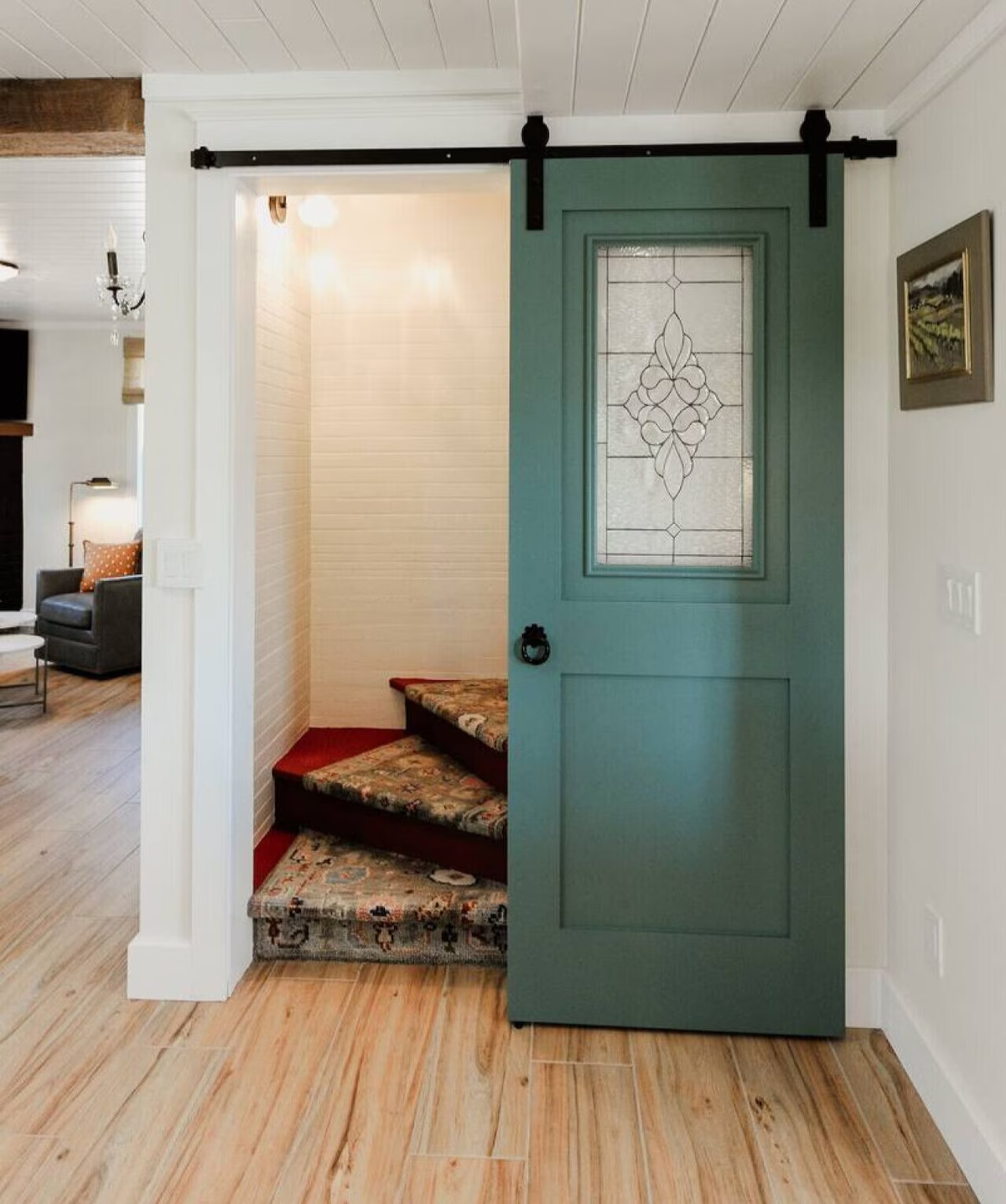 A turquoise sliding barn door with glass panel, open to reveal a small staircase with a floral rug. The room has light-colored walls, wood flooring, and a framed painting on the wall.