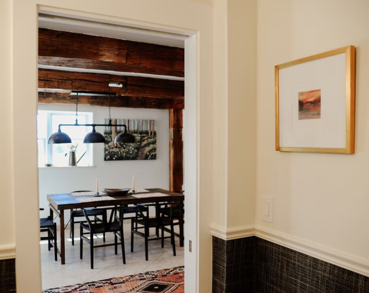 View of a dining room with a wooden table, black chairs, hanging black pendant lights, and artwork on the wall. The doorway frames the scene.