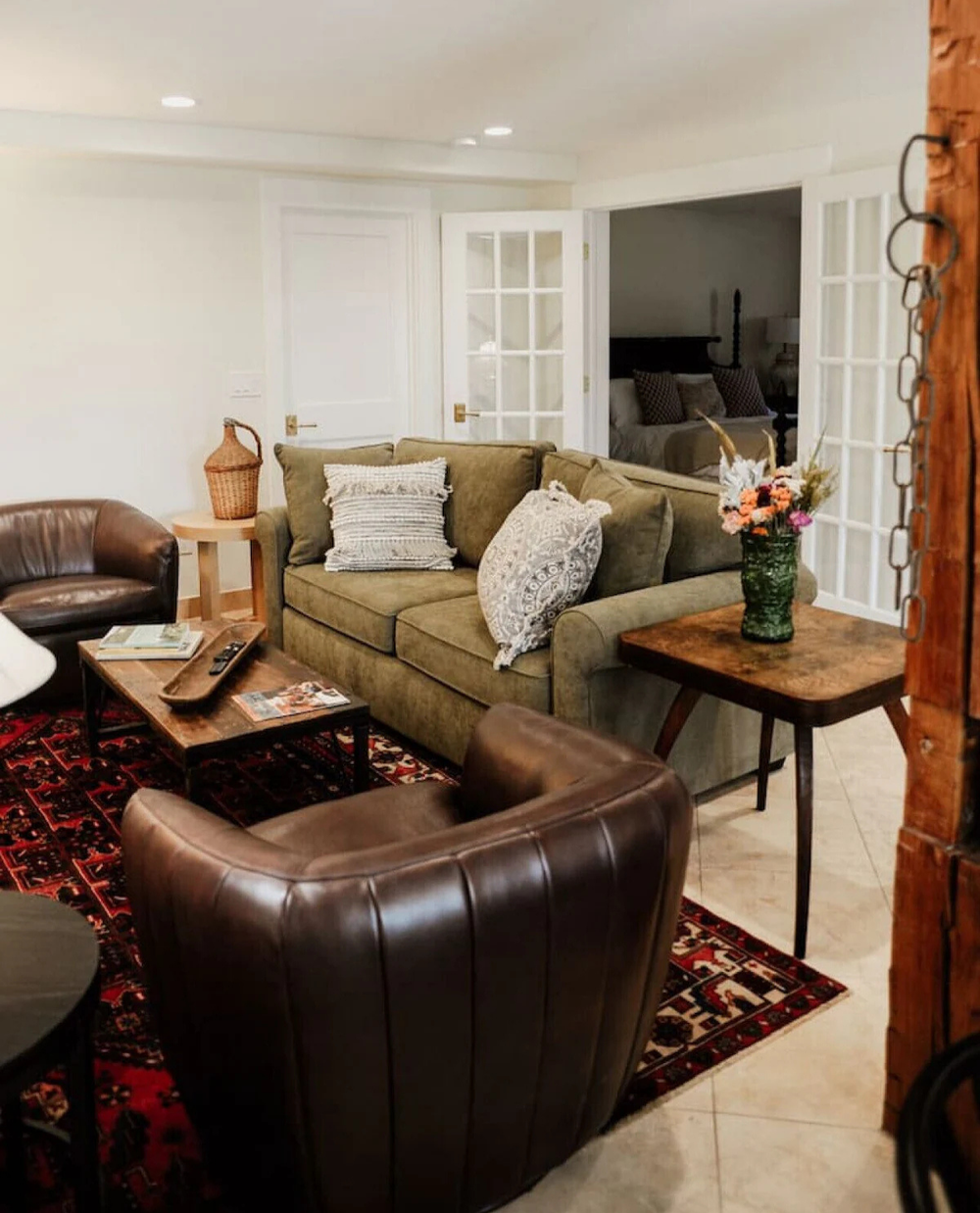 Living room with a green couch and patterned pillows, leading to a bedroom with a wooden bed frame and wall art, with glass double doors and a partial view of a dining area with a floral arrangement.