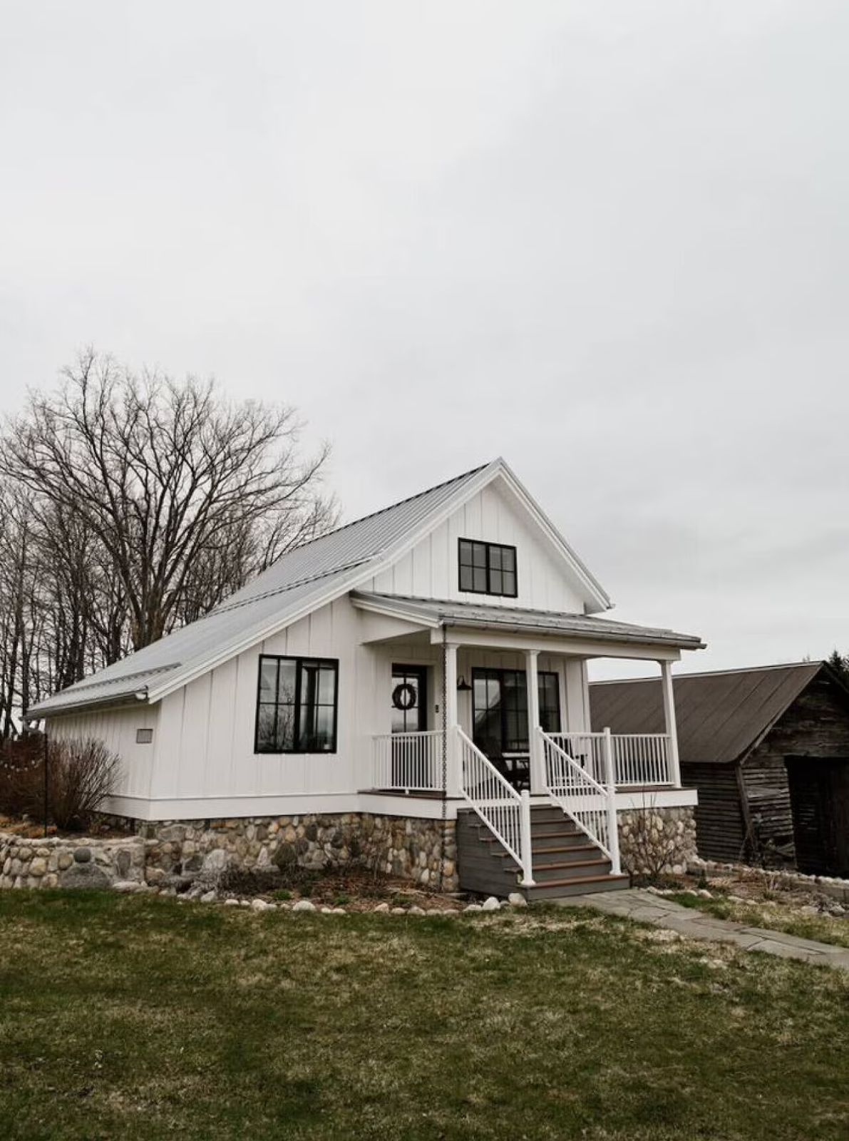 White house with black window frames and a stone foundation, elevated with stairs leading to porch, next to an old wooden shed in a yard with trees in the background.