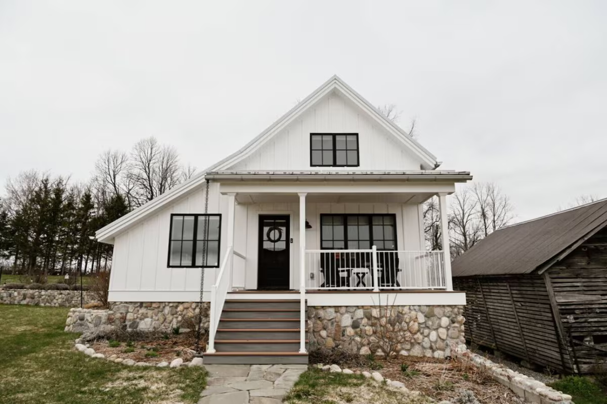 White two-story house with front porch, black door, stone foundation, and black window frames, surrounded by a garden and trees, under a cloudy sky.