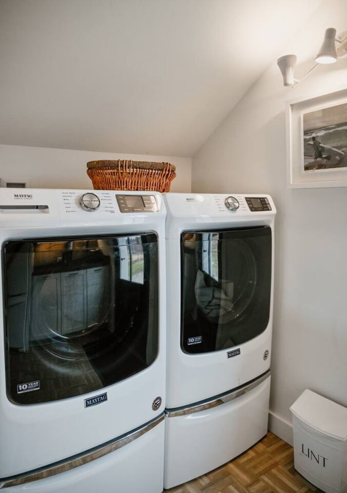 A laundry room with a white washer and dryer side by side, basket on top, framed picture on wall, and a LINT container nearby.