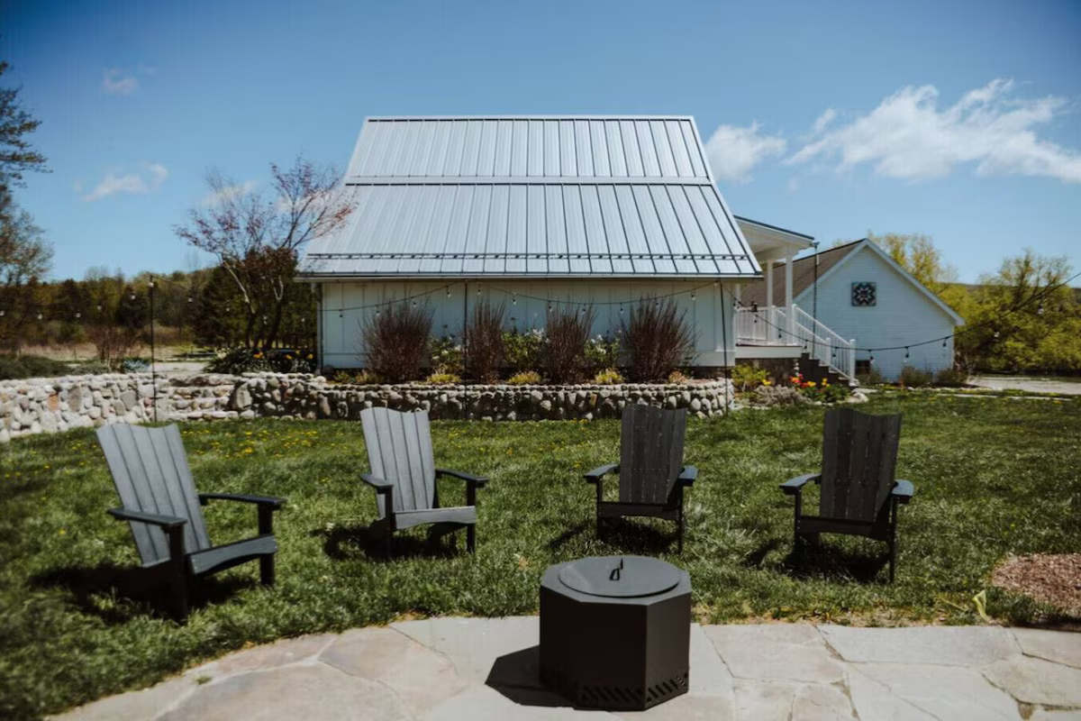 Outdoor backyard scene with four Adirondack chairs arranged in a semi-circle around a fire pit on a grassy lawn, with a white house with a metal roof and stone landscaping in the background, under a blue sky with clouds.