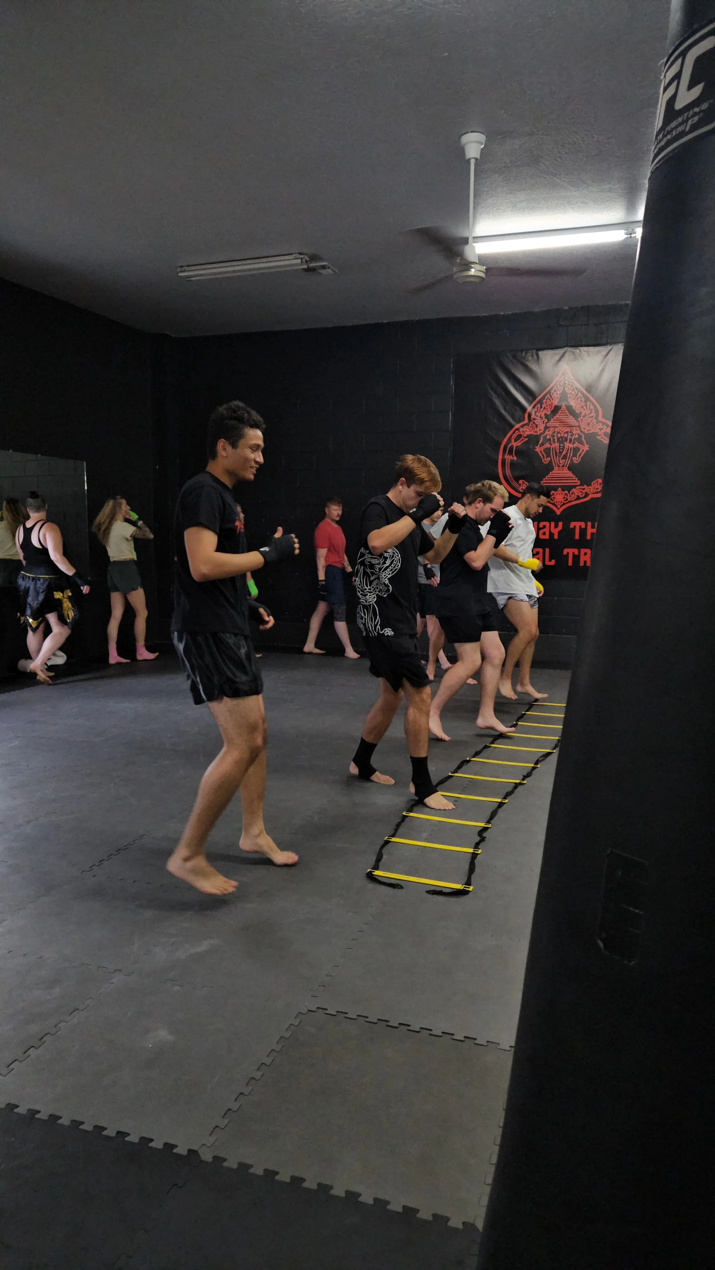 A group of martial arts students practicing agility ladder drills in a black-walled training center with Thai and MMA training branding.