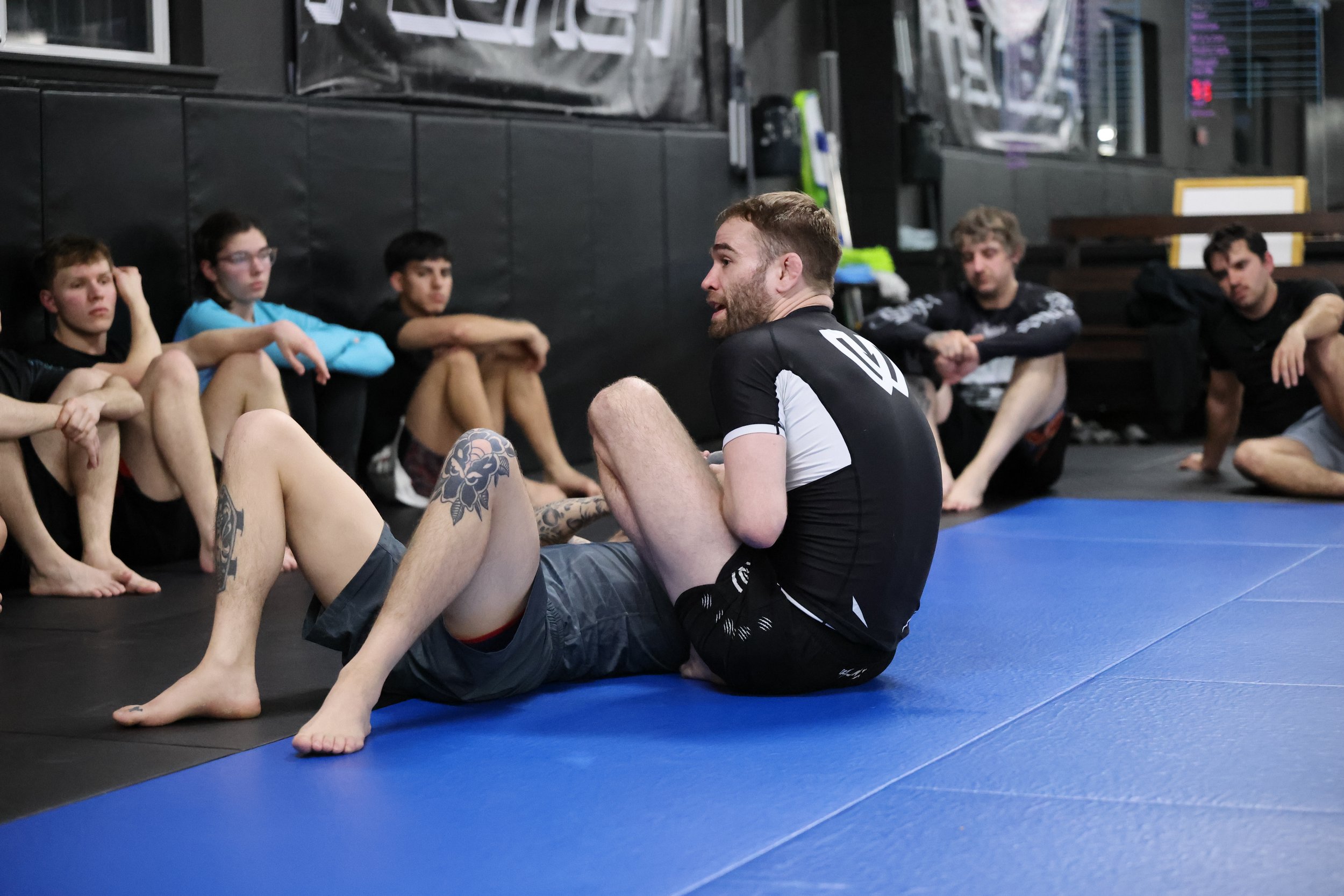 Martial arts students practicing Brazilian Jiu-Jitsu on mats in a gym, with some seated and others standing, while some are performing ground techniques.