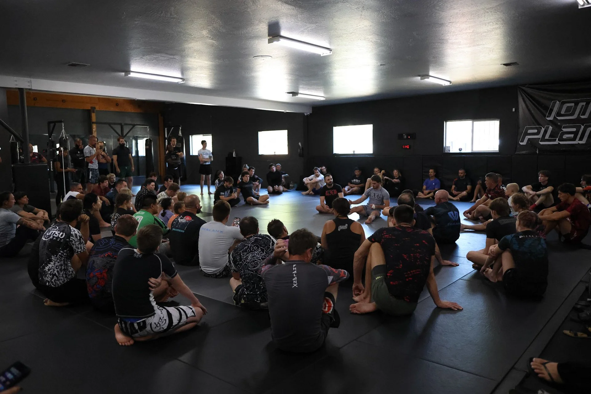 Martial arts students practicing Brazilian Jiu-Jitsu on mats in a gym, with some seated and others standing, while some are performing ground techniques.