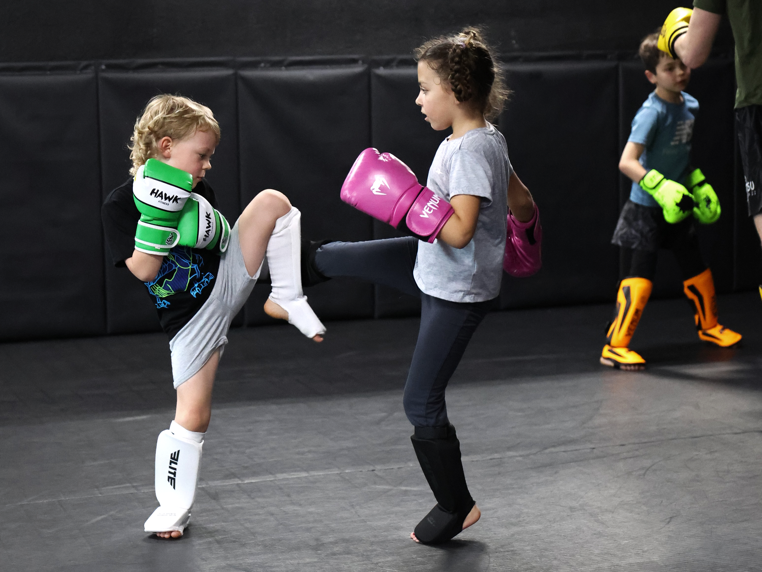 Kids and an instructor practicing martial arts at a martial arts gym with punching bags and a mirrored wall.