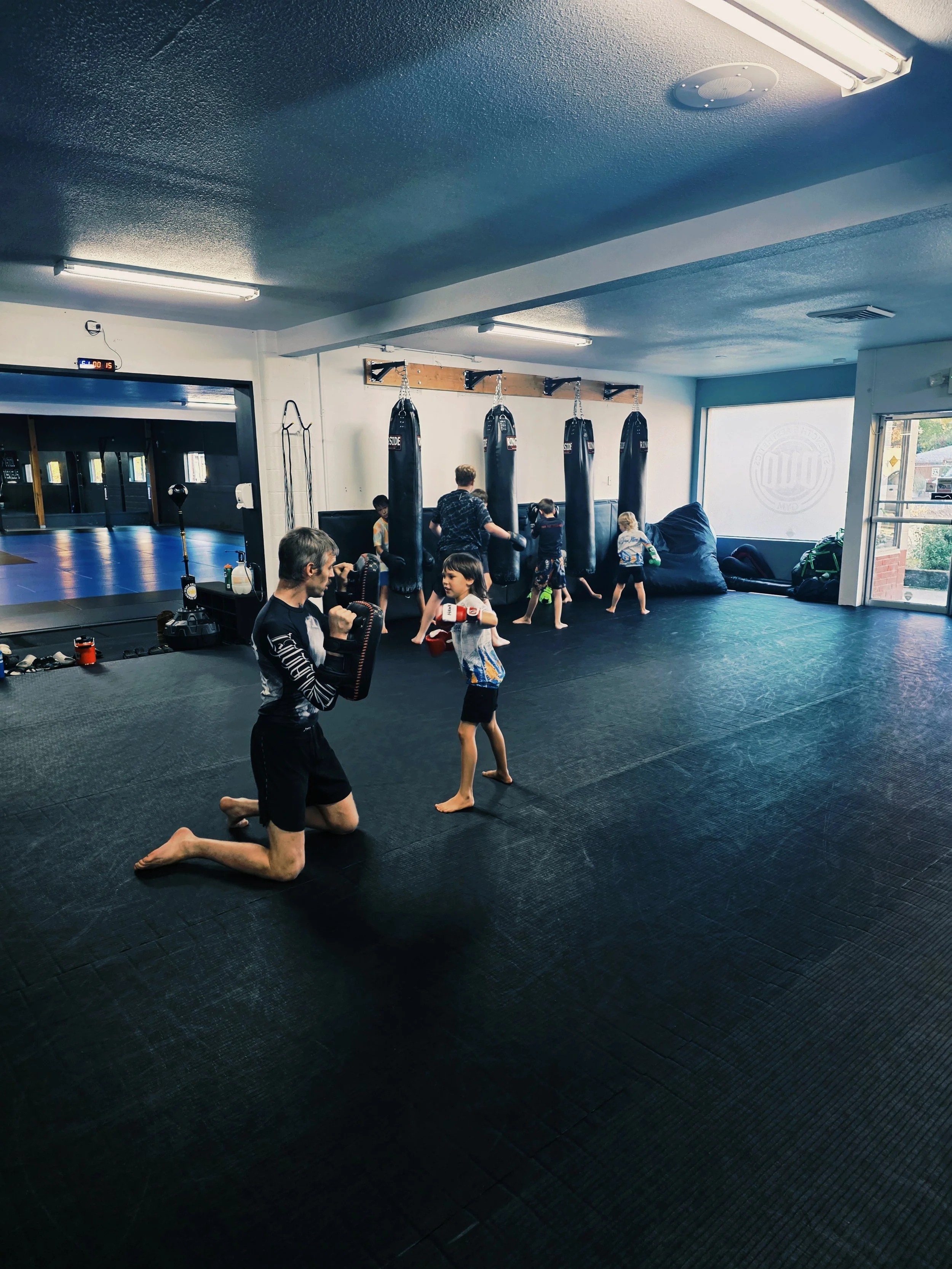 Kids and an instructor practicing martial arts at a martial arts gym with punching bags and a mirrored wall.