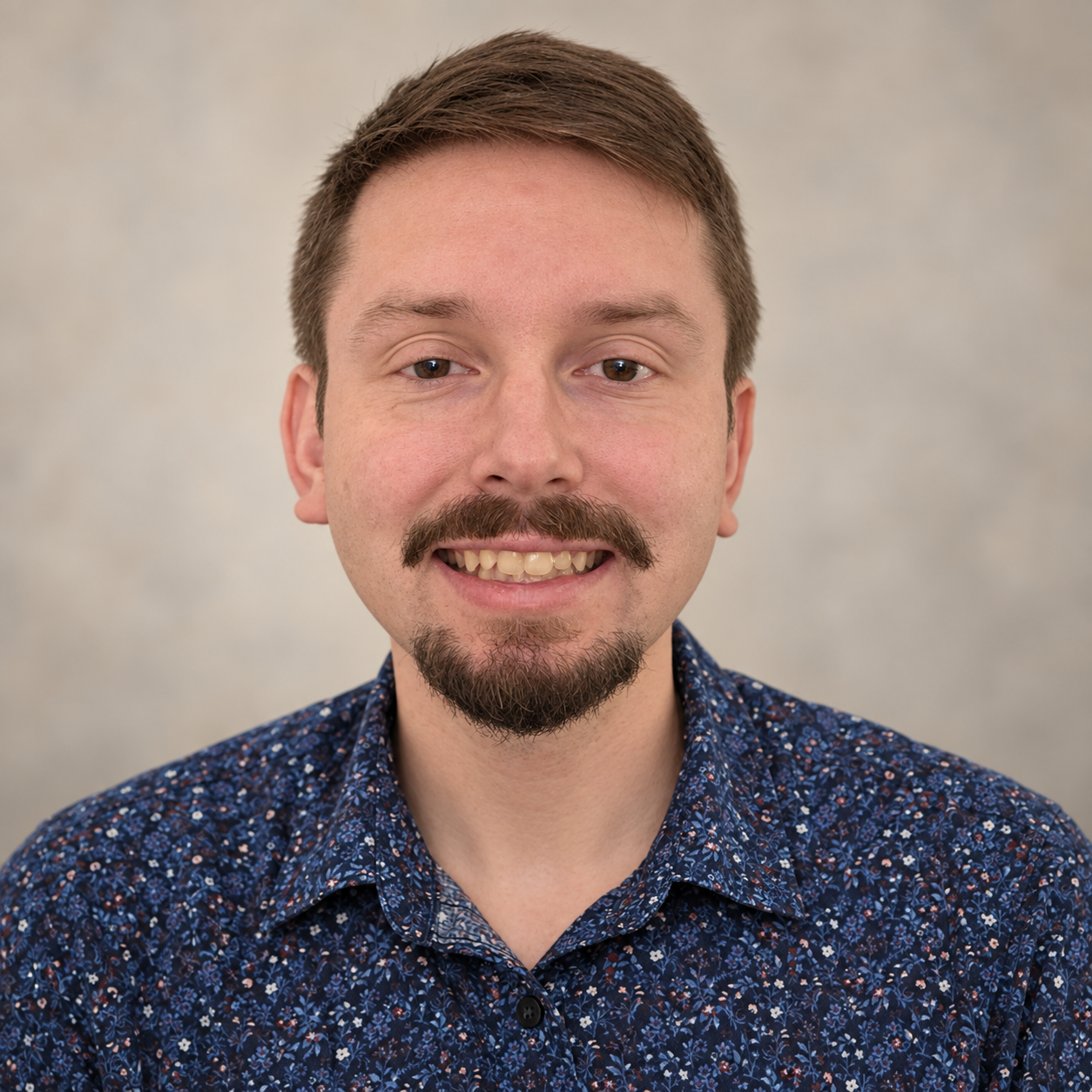 Headshot of a smiling young man with brown hair, a mustache, and goatee, wearing a blue floral shirt, against a plain background.