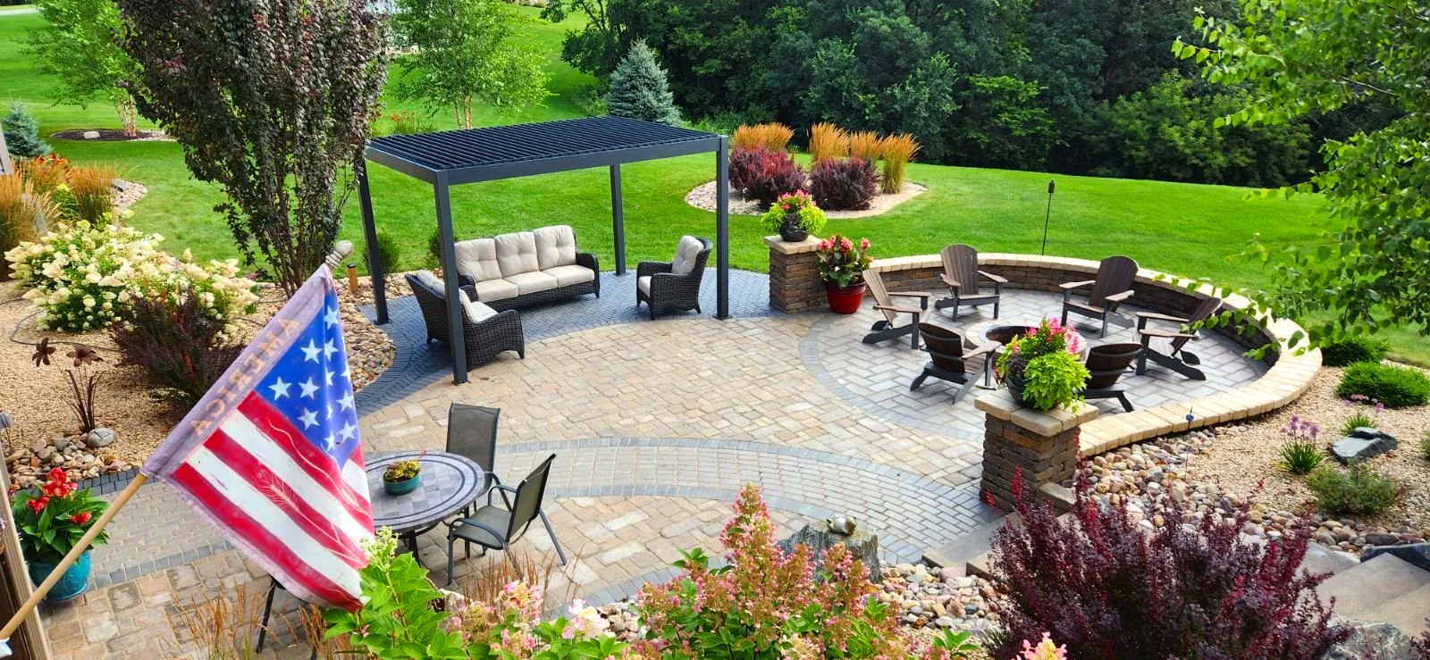 A backyard patio with seating areas, including a round table with chairs, a covered sofa set, and a circular fire pit with Adirondack chairs around it. The patio is surrounded by landscaped plants, flowers, and trees, with a grassy lawn in the backgr