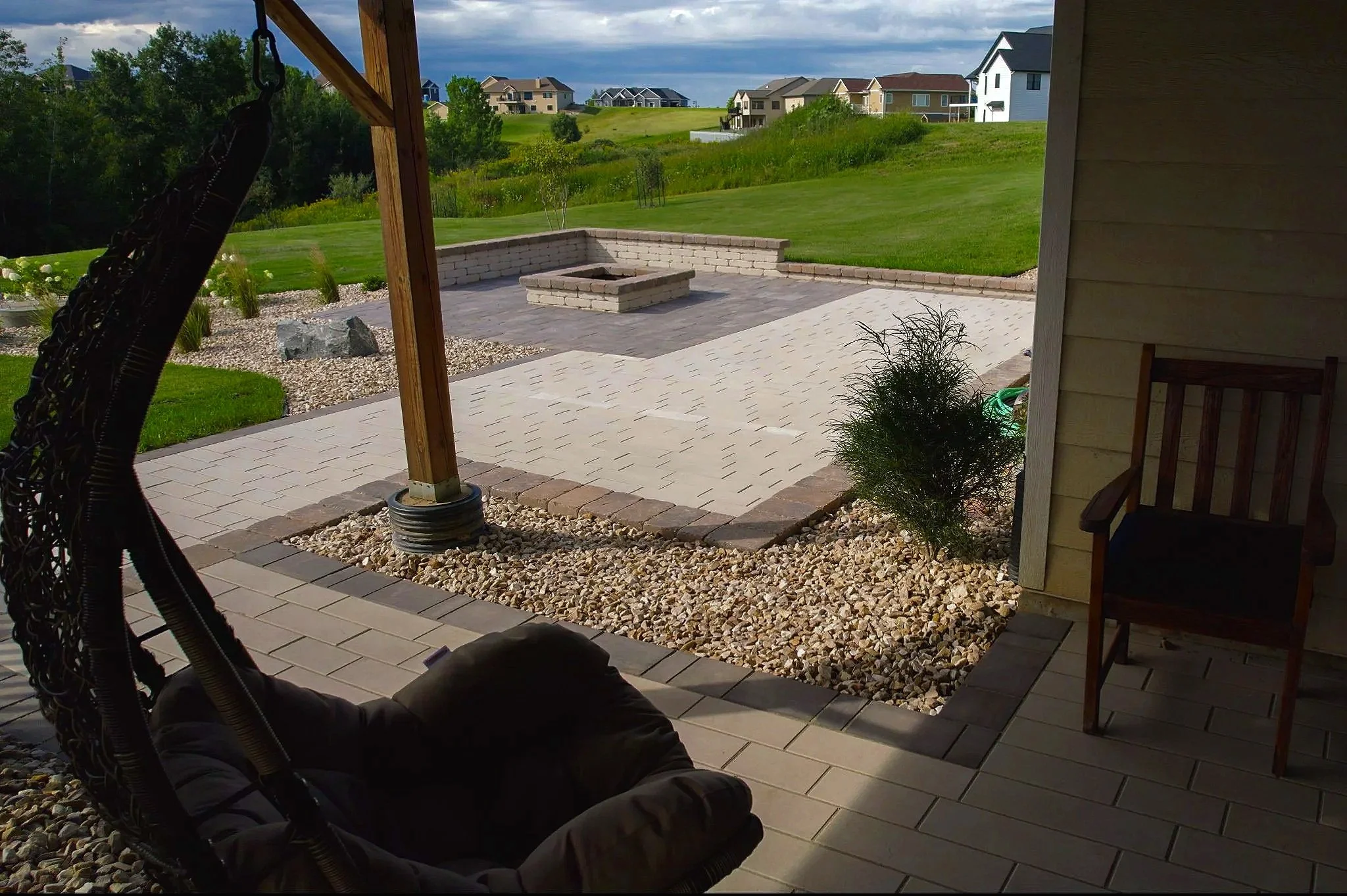 View from a covered patio overlooking a backyard with a stone paved area, a fire pit, and well-maintained lawn and houses in the distance. There is a hanging chair on the patio and a wooden chair against the wall.
