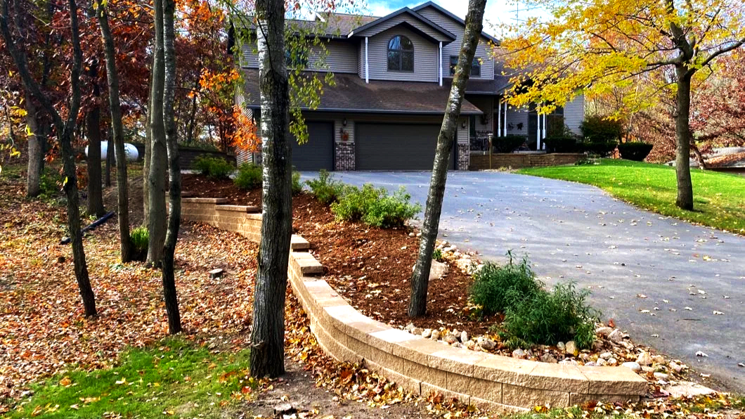 A two-story gray house with a dark roof and attached garage, surrounded by trees with autumn leaves, and a curved driveway leading up to the garage.