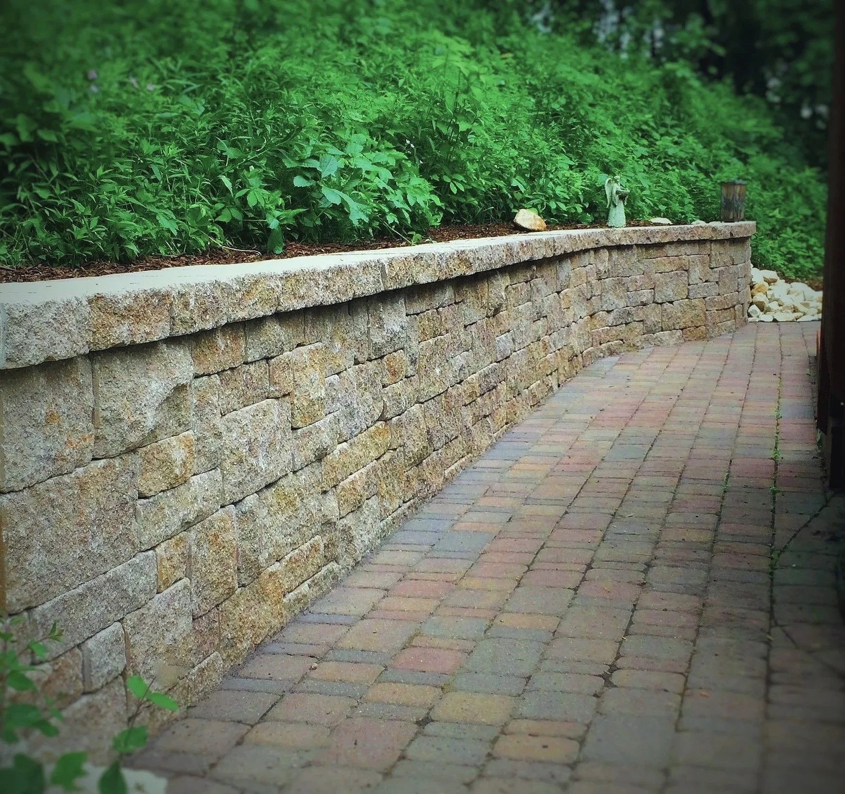 A stone retaining wall with a concrete cap, topped with green foliage, along a paved brick pathway.