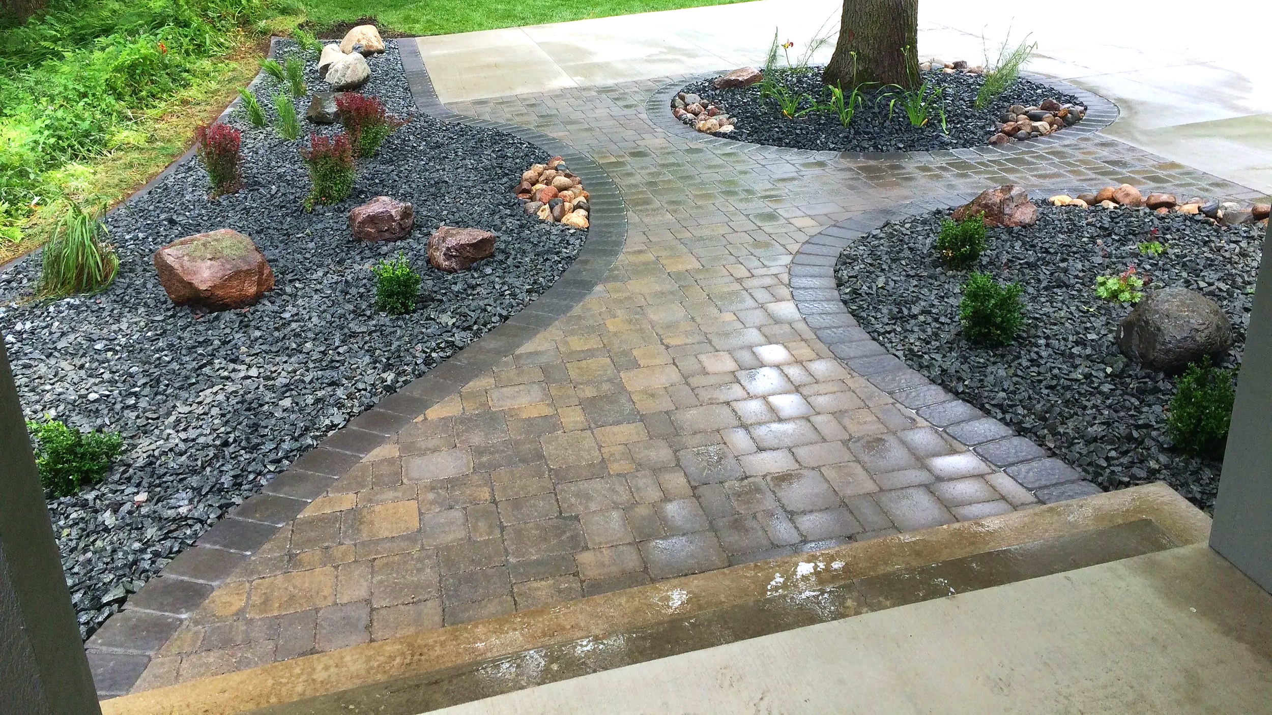 A landscaped backyard with a curved stone pathway, a small garden bed with rocks and plants around a large tree, and a grassy area with a driveway and a pickup truck in the background.