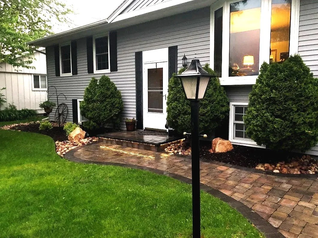 Front yard of a house with a paved walkway, trimmed bushes, and a outdoor lamp post, during dusk with warm lighting visible through the window.