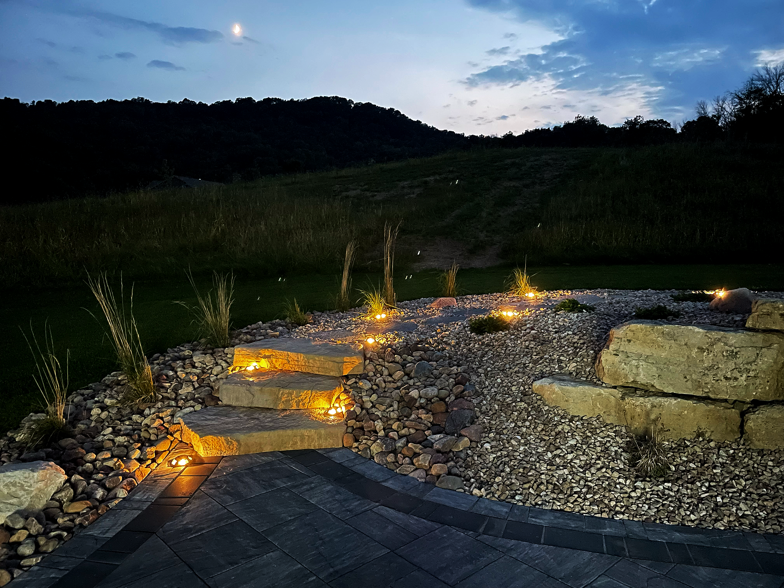 A landscaped yard at dusk with stone steps, decorative rocks, plants, and small lights illuminating the area, with a grassy hill and trees in the background under a twilight sky.