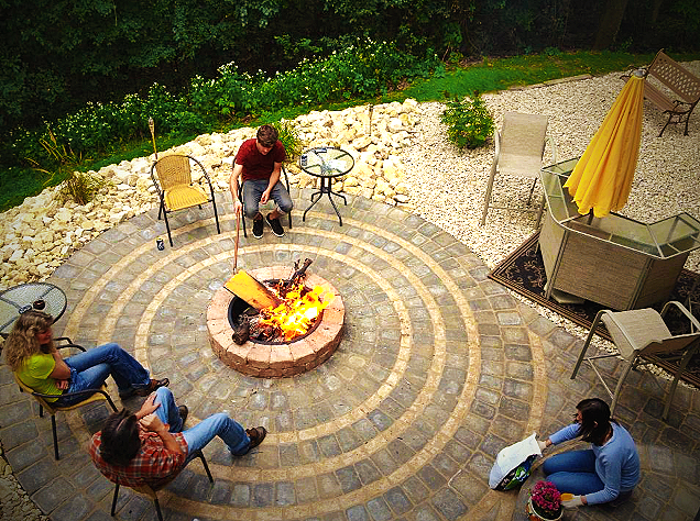 Four people sitting around a fire pit on a circular patio with outdoor furniture and a garden in the background.