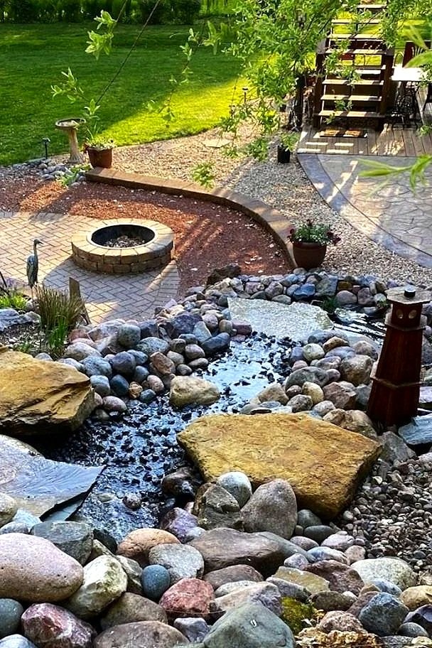 A landscaped backyard featuring a small stream with rocks, a fire pit surrounded by a paved area, potted plants, a wooden deck with stairs, and a grassy lawn with trees in the background.