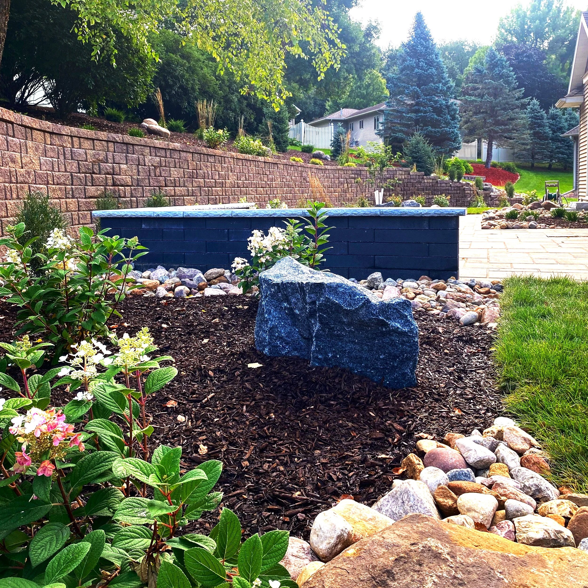 A landscaped backyard garden with a stone border, mulch bed, and flowering plants, featuring a large rock and a blue-painted brick wall, with trees and a house in the background.
