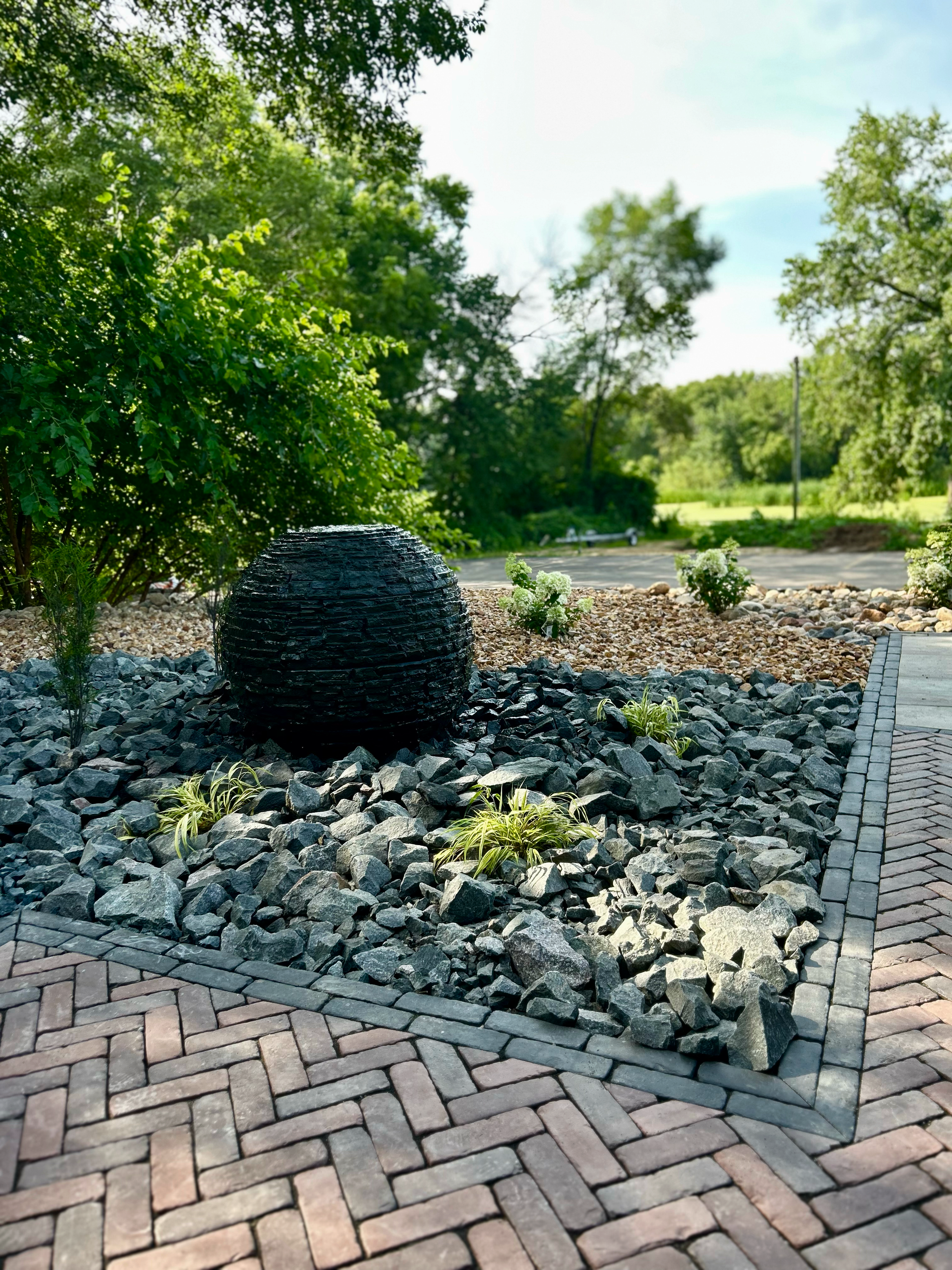 Decorative outdoor garden with a large black spherical fountain, surrounded by rocks, small green plants, and bricks with a landscaped background of trees and shrubs.