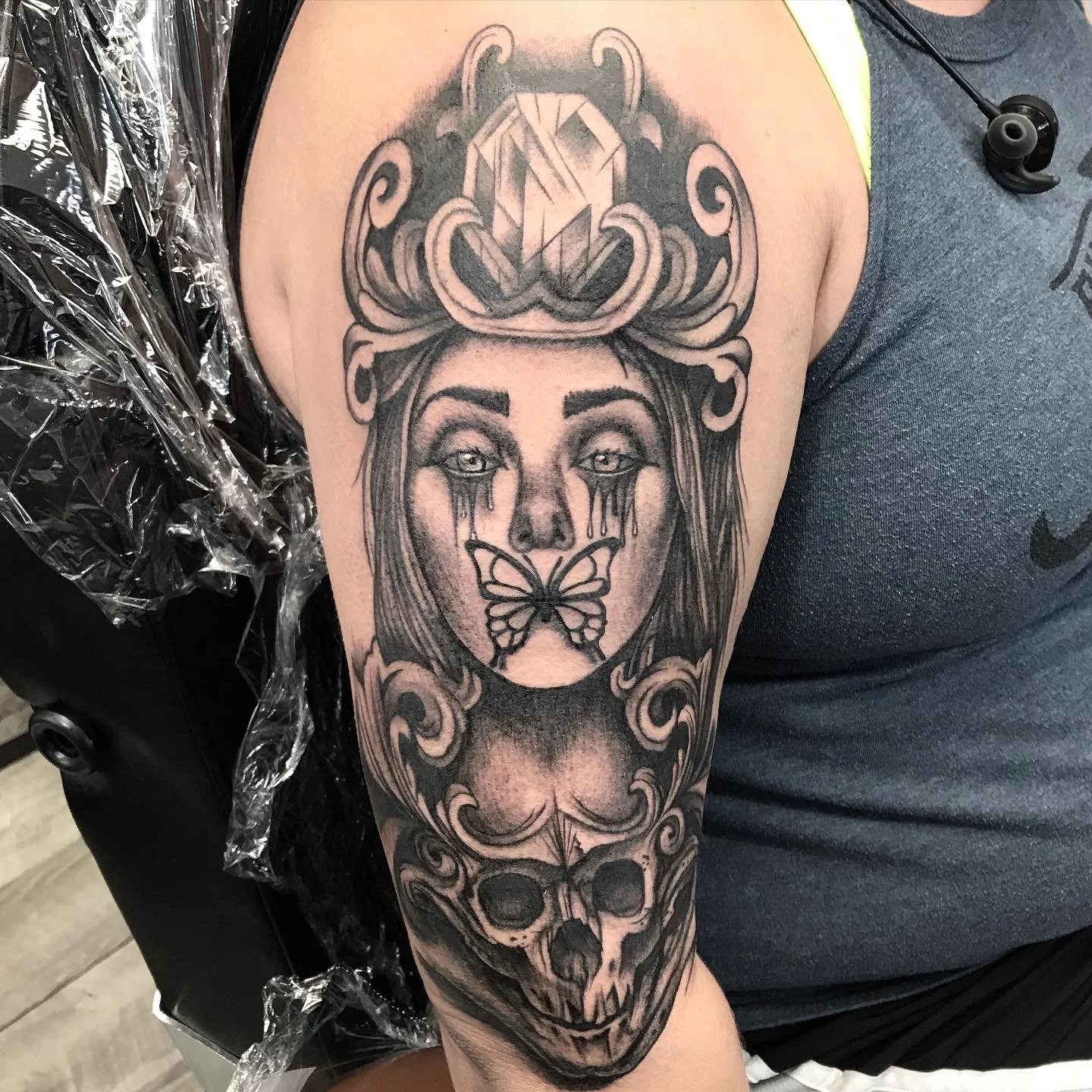 A black and gray tattoo on a person's arm featuring a woman's face with tears, a butterfly near her mouth, a skull at the bottom, and ornamental designs around the face and skull.